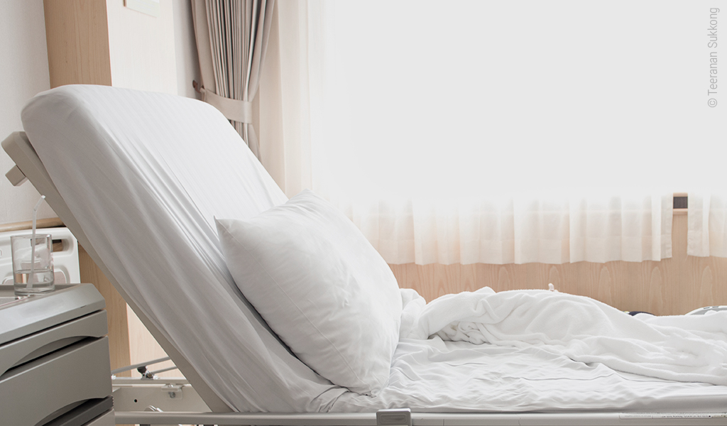 Foto: Ein Pflegebett mit aufgerichtetem Kopfteil; daneben steht ein Nachtkasten, auf dem ein Wasserglas mit Strohhalm steht.