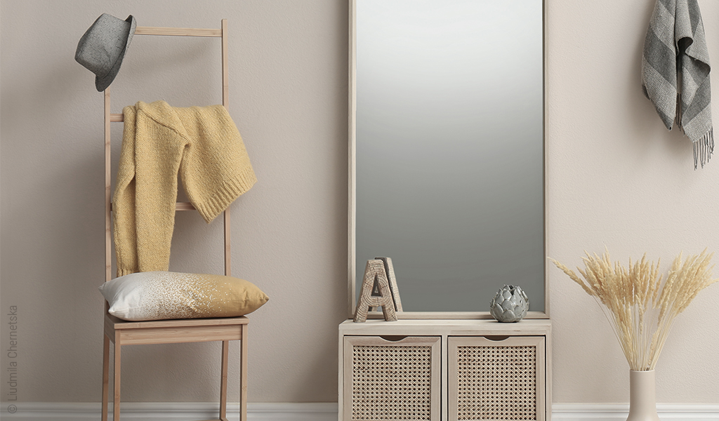 Foto: An der Wand eines beige eingerichteten Zimmers steht ein Kleiderstuhl, ein Spiegel auf einer Kommode, eine Blumenvase mit getrocknetem Getreide auf dem Boden und an der Wand hängt ein Schal.