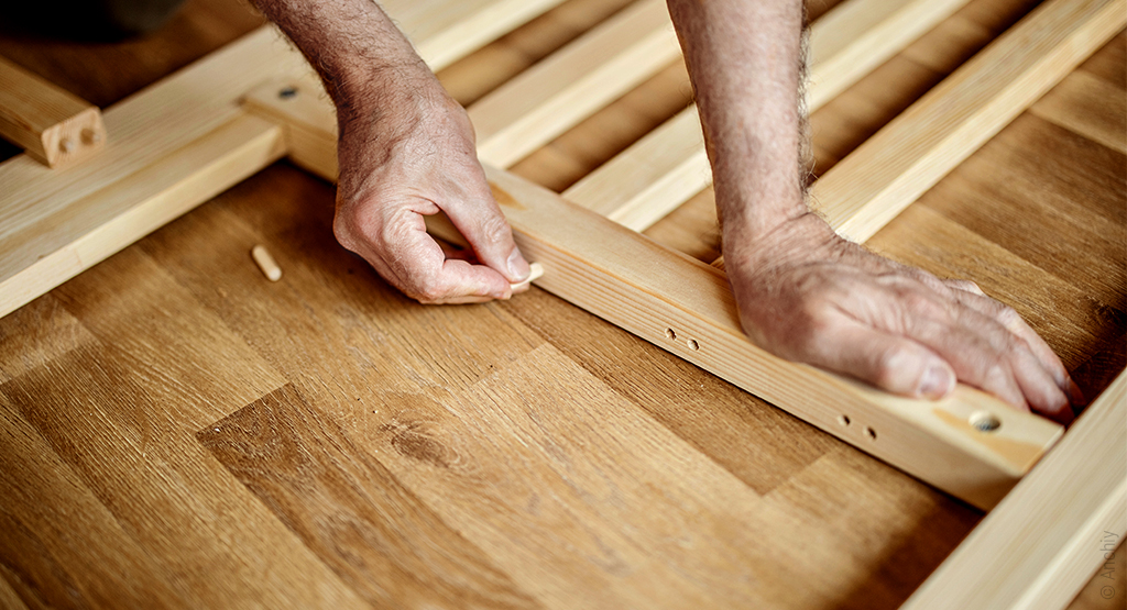 Foto: Teils verschraubte teils lose Holzlatten auf dem Boden. Eine Hand stützt sich auf eine Leiste, eine andere steckt einen Holzdübel in eine Bohrung.