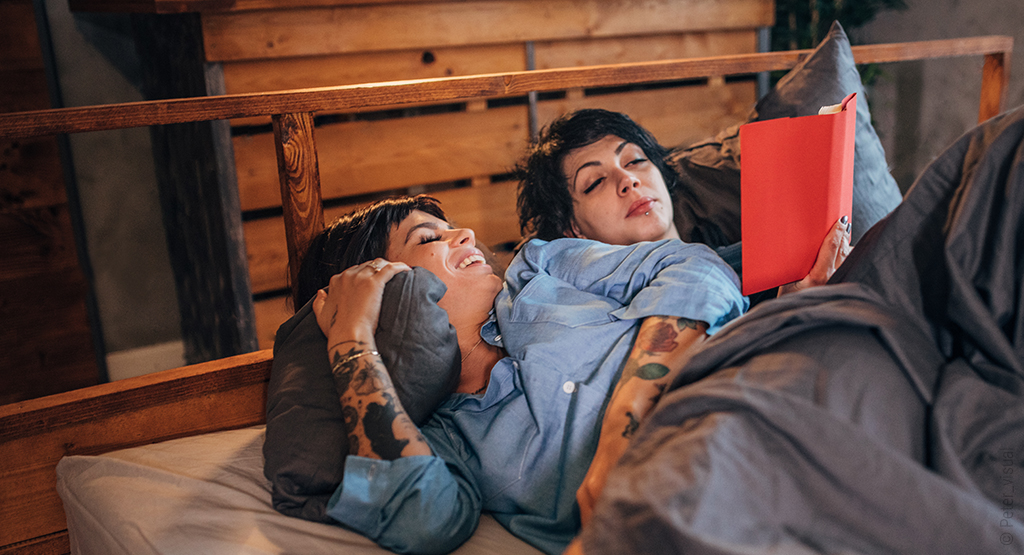 Foto: Zwei Personen liegen in einem Holzbett und sehen in das gleiche Buch