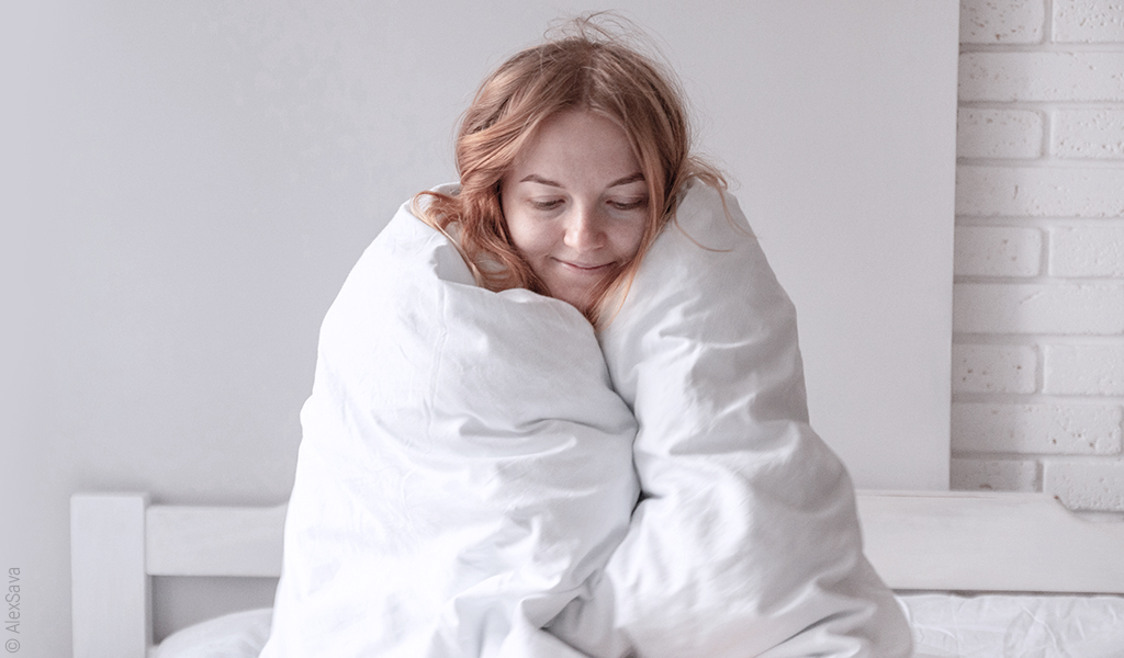 Foto: Eine Person sitzt aufrecht in eine dicke Bettdecke gewickelt in einem Bett.