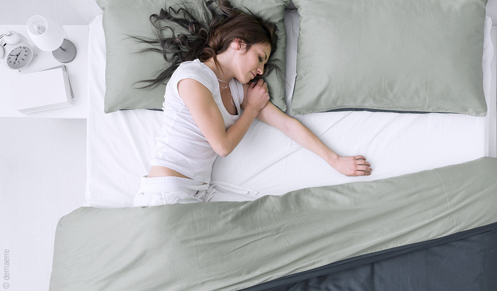 Foto in Vogelperspektive: Eine Person in weißem Schlafanzug liegt ausgestreckt allein in einem Doppelbett.