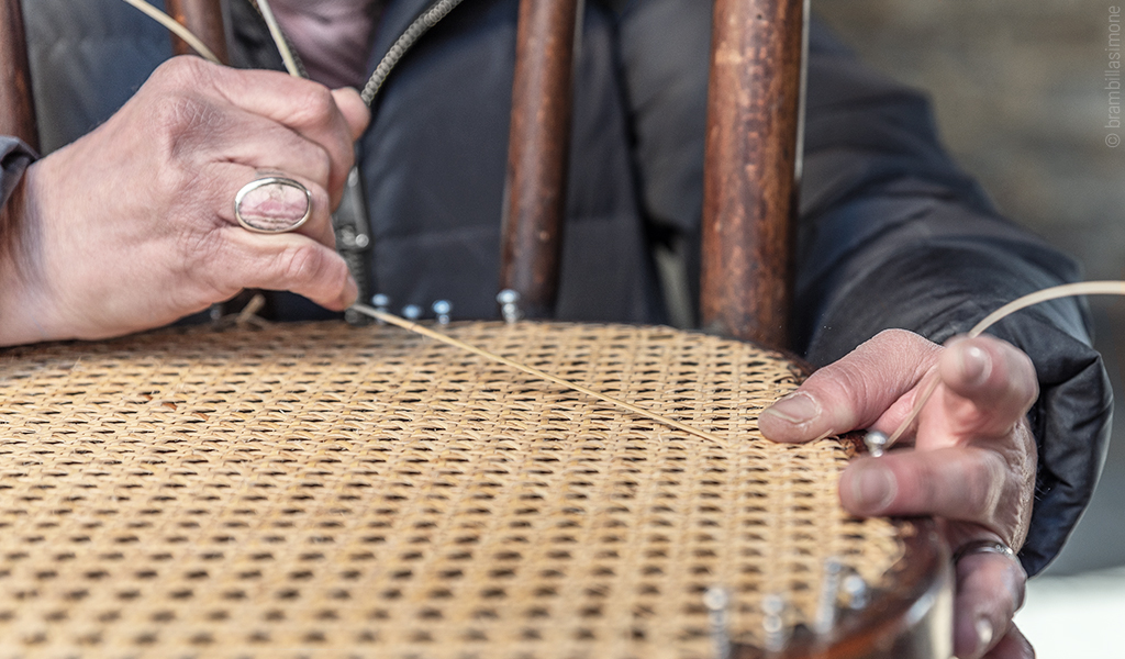 Foto: Nahaufnahme von zwei Händen, die an der Sizfläche eines Rattanstuhls arbeiten.