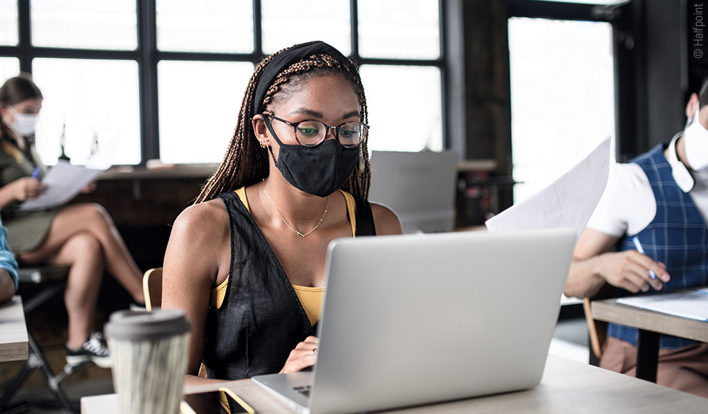 Foto: Ein Großraumbüro. An Tischen sitzen Mitarbeitende auf Abstand mit Laptop und FFP2-Maske.