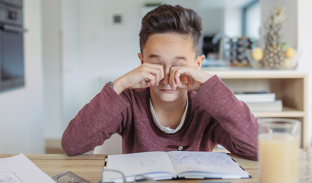 Foto: Eine Person sitzt an einem Schreibtisch und reibt sich die Augen.