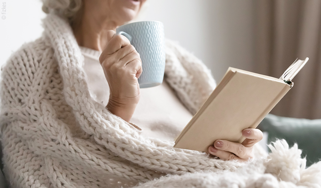 Foto: Eine Person sitzt eingewickelt in eine Decke auf einer Couch. Sie hat ein Buch in der einen Hand, eine Tasse in der anderen Hand.