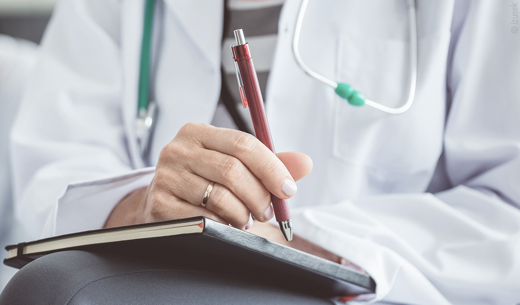 Foto: Eine Person mit Kugelschreiber in der Hand, ein Notizbuch auf dem Schoß