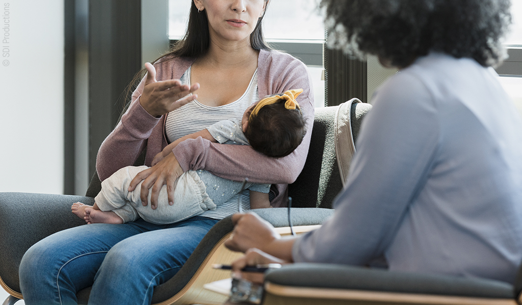 Foto: Eine Person mit Baby auf dem Arm im Gespräch mit einer weiteren Person, die sich dazu Notizen macht. 