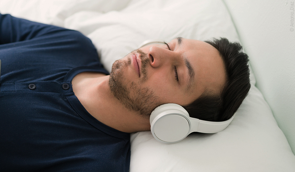 Foto: Eine Person liegt in Rückenlage auf dem Bett und hat dabei Kopfhörer auf. 