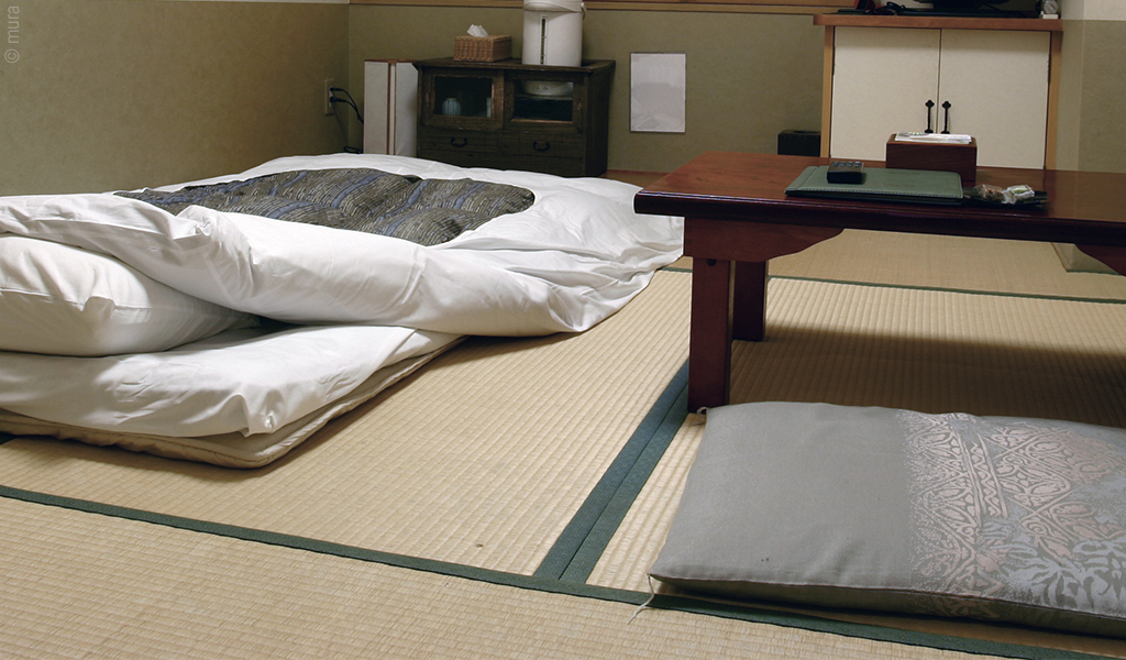 Foto: In einem Zimmer, dessen Boden mit Reisstrohmatten ausgekleidet ist, liegt links ein Futon und rechts steht ein niedriger Tisch mit Sitzkissen davor.