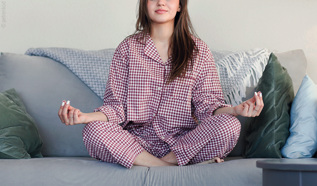 Foto: Eine Person sitzt im Pyjama auf einer Couch in einer Meditationspose.