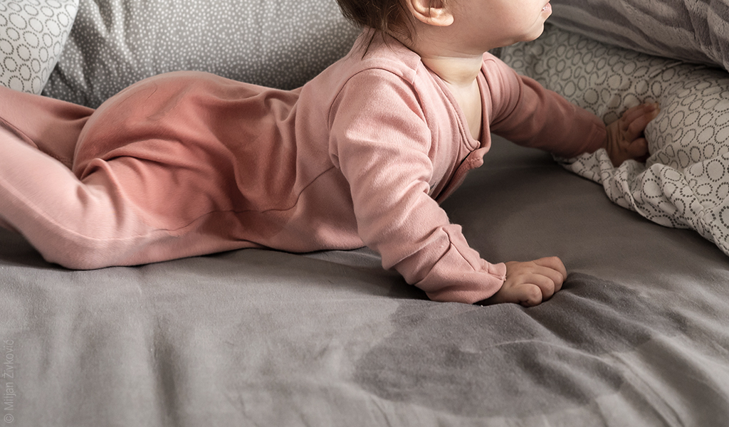 Foto: Ein Baby liegt den Oberkörper aufgerichtet zur Krabbelposition in einem Bett. Auf dem Laken ist ein großer nasser Fleck.
