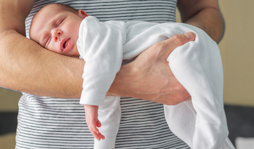 Foto: Eine Person hält ein schlafendes Baby im Fliegergriff.