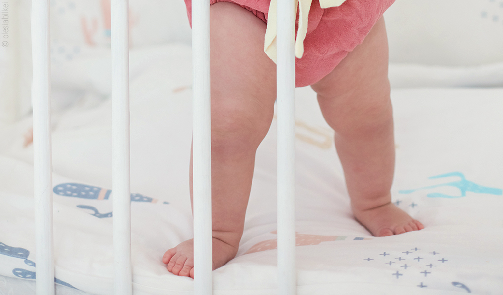 Foto: Hinter einem Bettgitter stehen zwei Babybeine auf einer Babymatratze.