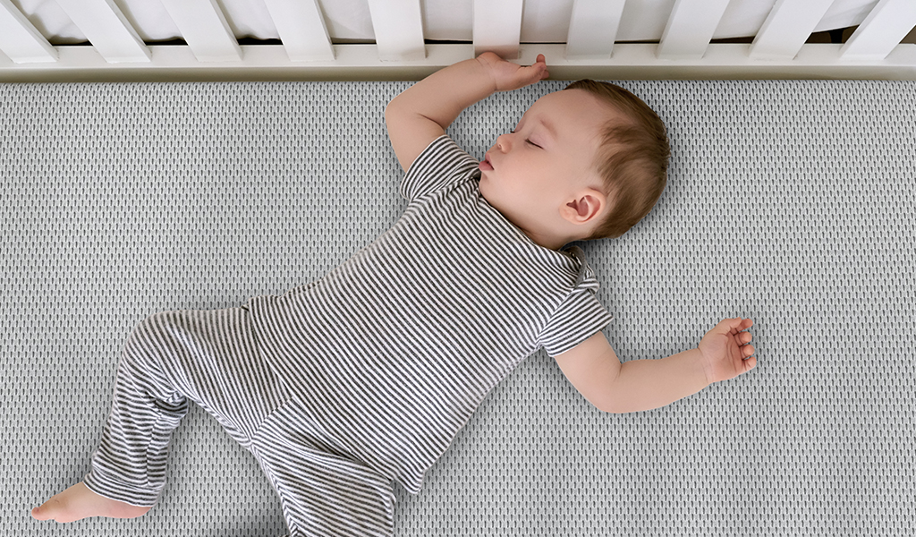 Foto: Ein Baby liegt auf dem Rücken in einem Kinderbett ohne Kissen oder Decke.