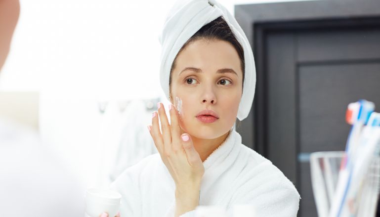 Mulher branca de roupão branco e toalha branca enrolada na cabeça, passando creme no rosto. Ela sabe a ordem da maquiagem.