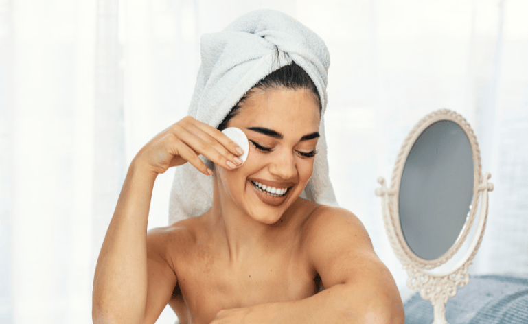 Mulher branca, enrolada em uma toalha branca de banho, na cabeça também, passando um produto no rosto com algodão e sorrindo.