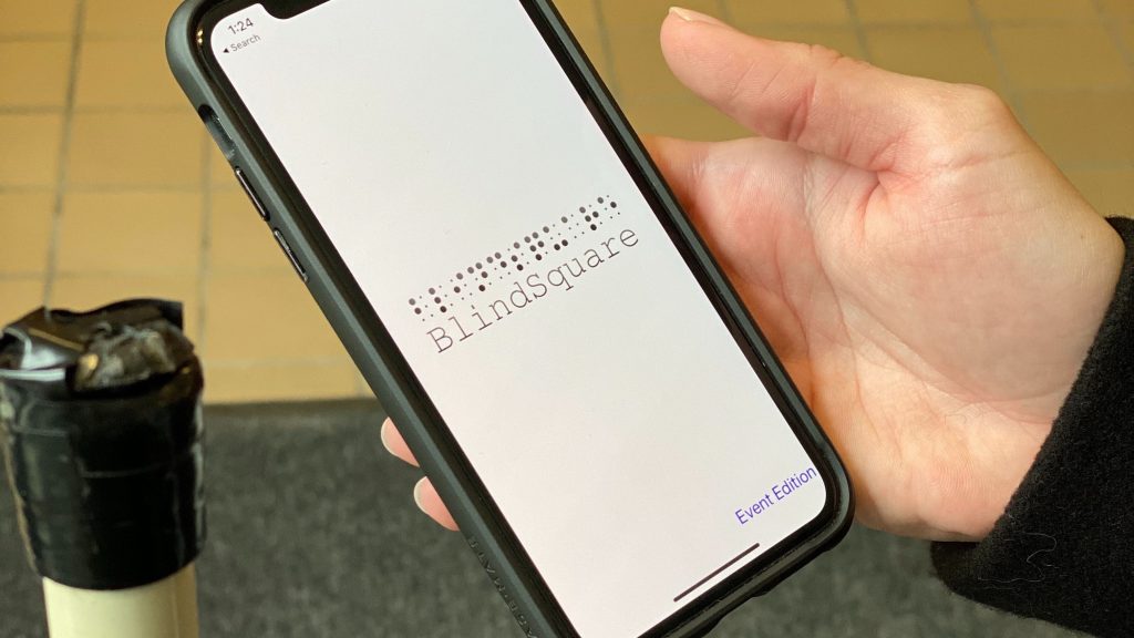 Image of someone holding a white cane and an iPhone with the BlindSquare Event app visible on the screen.