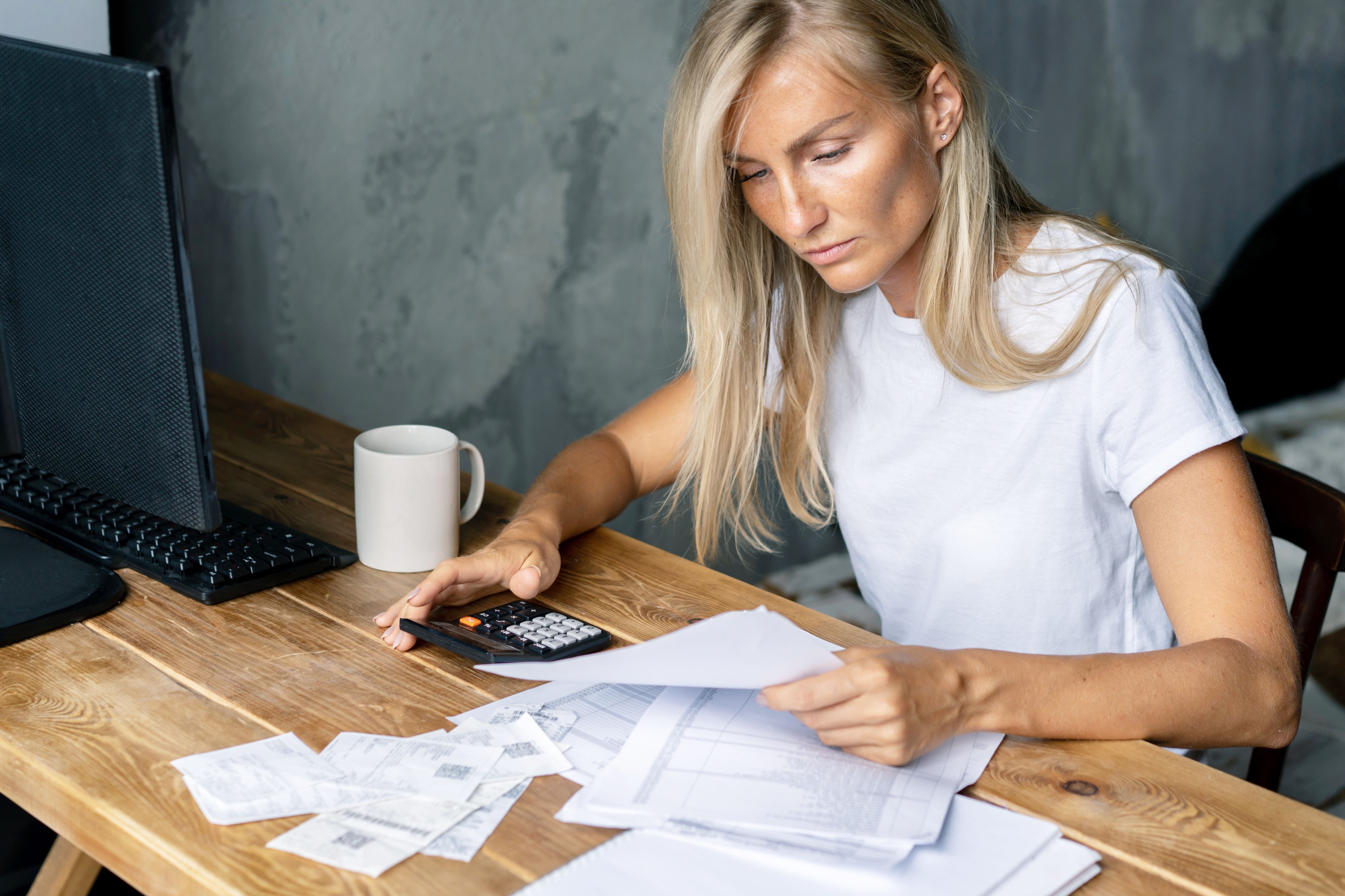 A blonde woman sits at a desk and works with bills and documents using a calculator