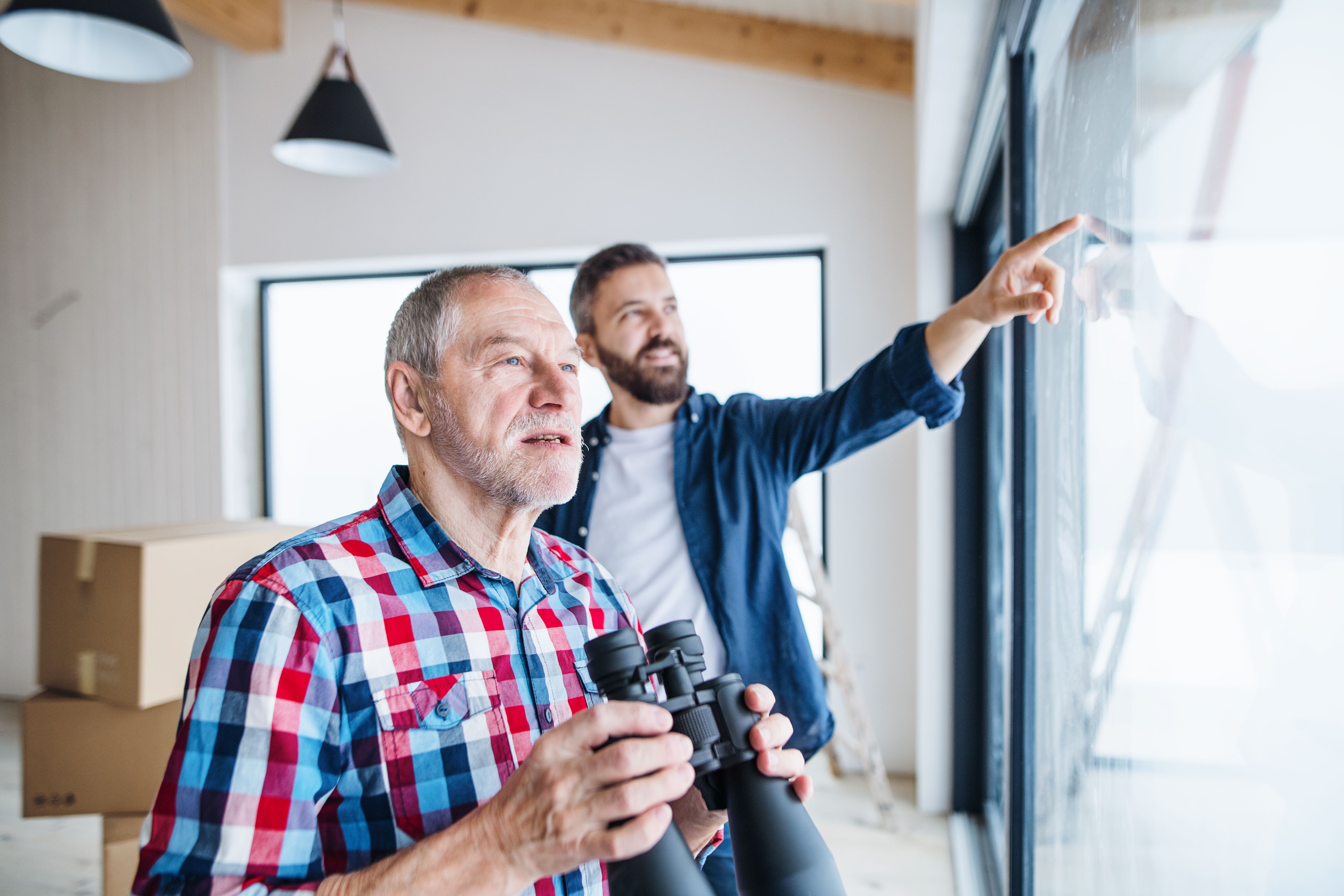 A senior man holding binoculars and his son inspecting a new apartment with boxes in the background 