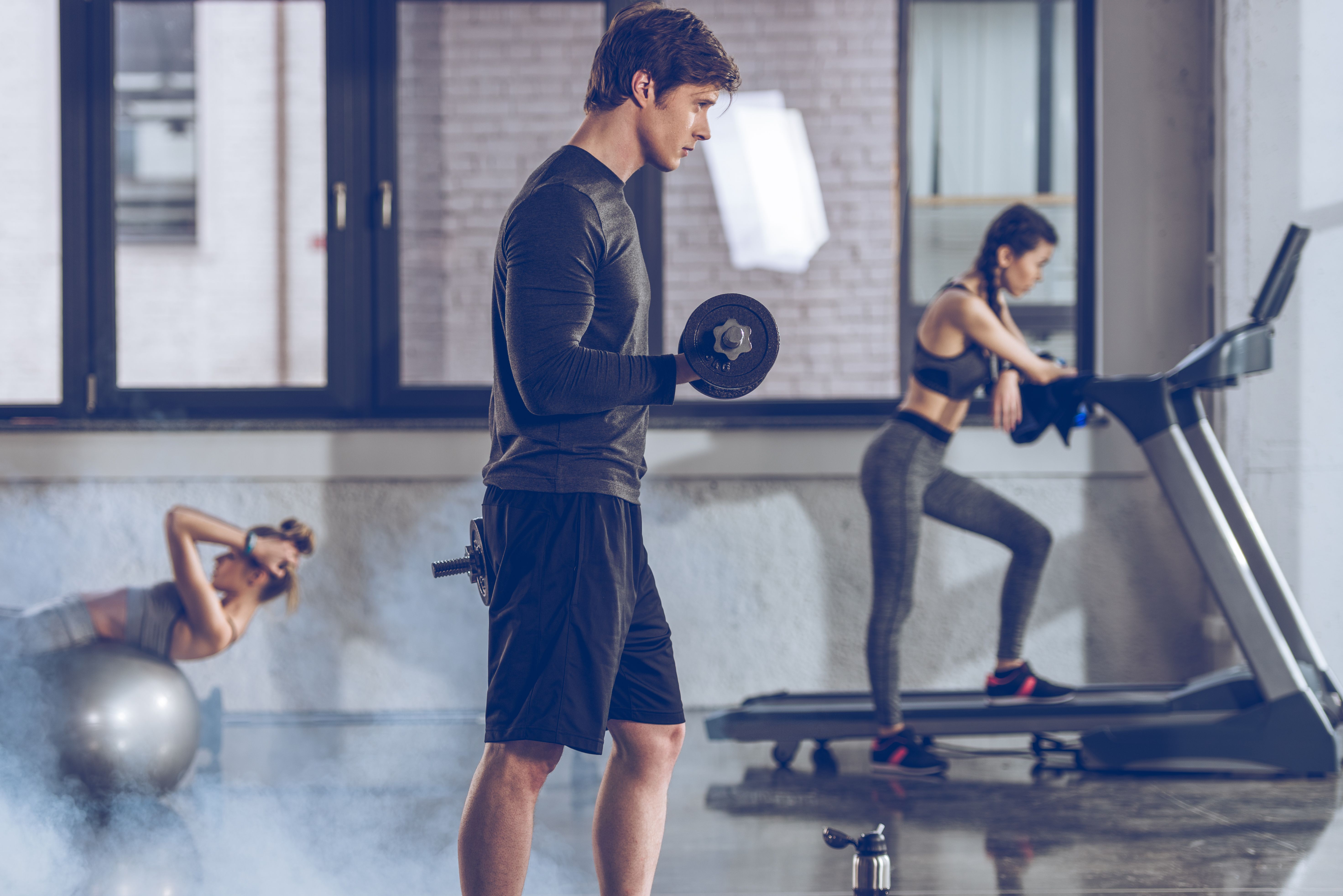 A young athletic man in a gym exercising with dumbbells with two women using fitness equipment in the background