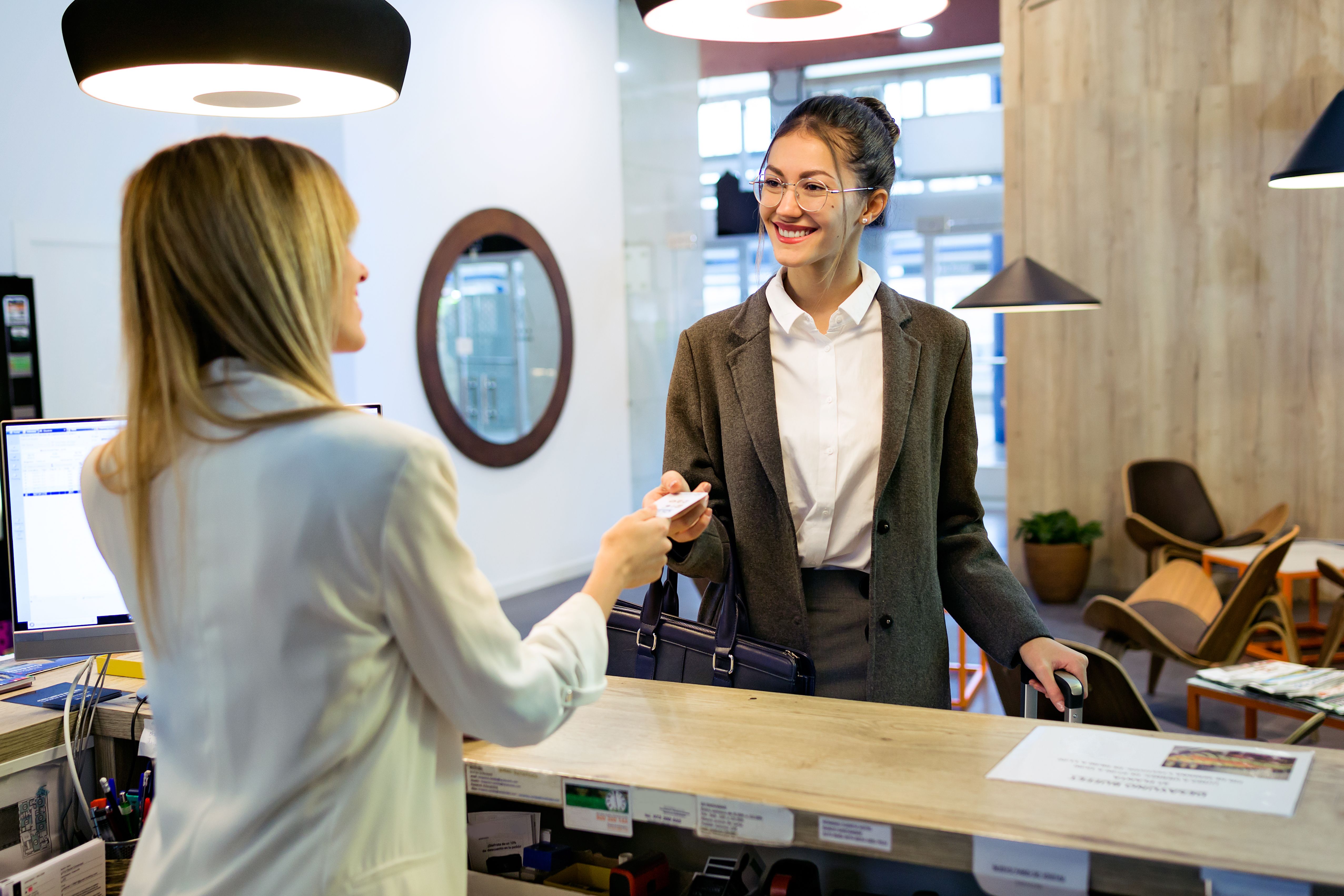 A businesswoman carrying luggage and talking to a building concierge with chairs, lights, and a mirror in the background 