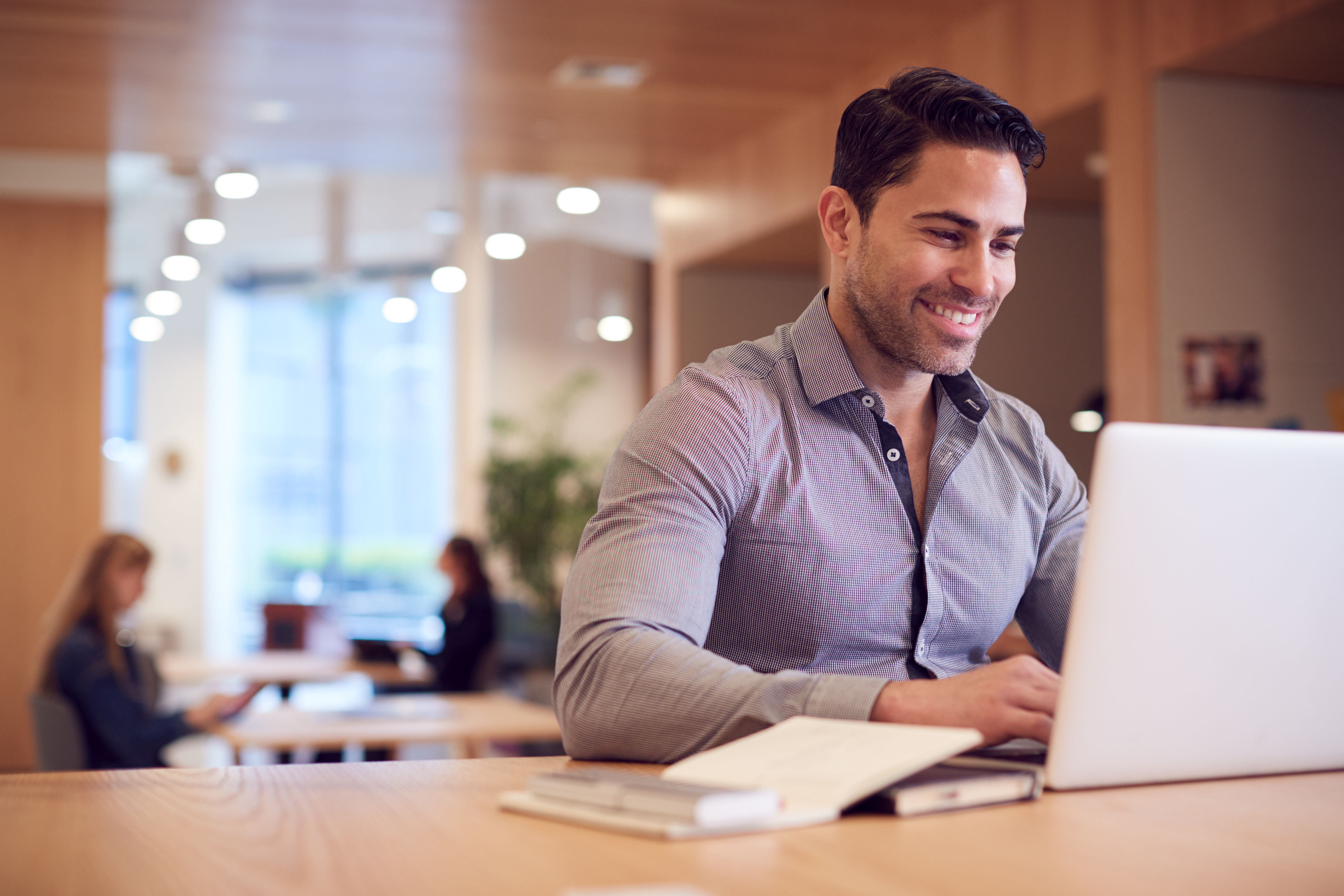 A smiling businessman using a laptop in a co-working space with two women in the background