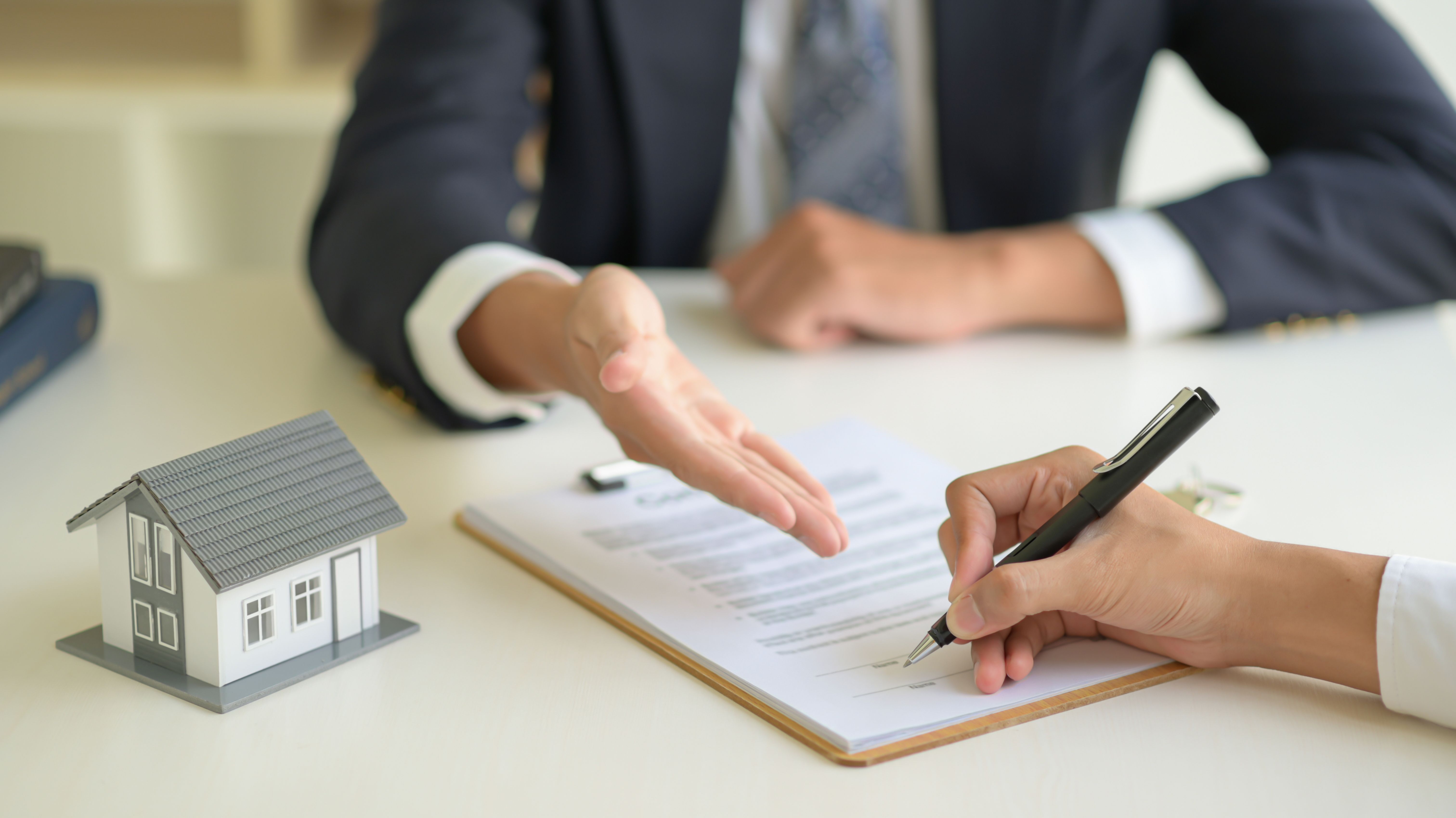 A tenant in a white shirt signing a property rental contract with their landlord in a black suit before moving in, next to a grey house model.