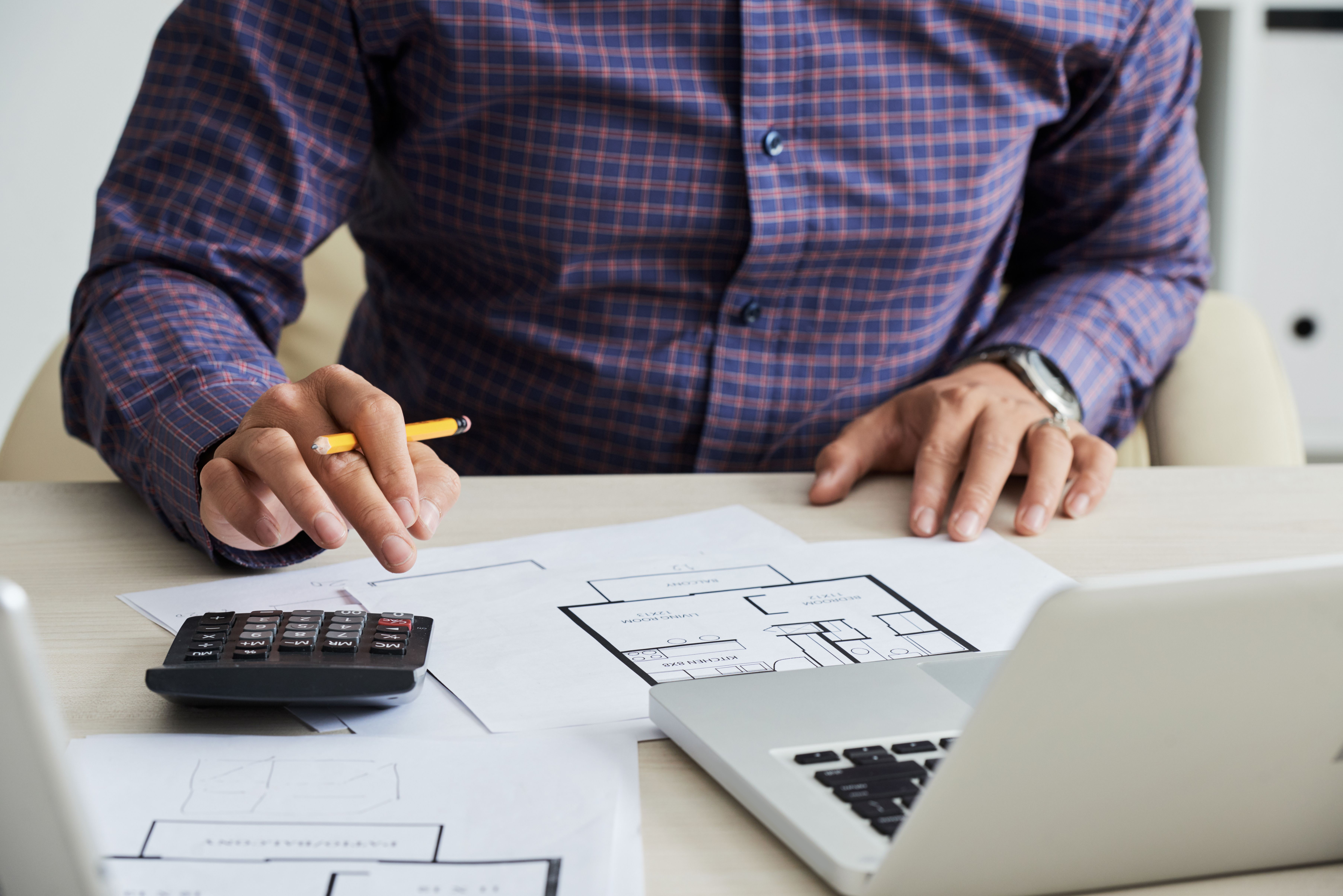 A landlord in a purple shirt carefully calculating his deposit deductions with a black calculator and a yellow pencil, next to a floorplan and a laptop.