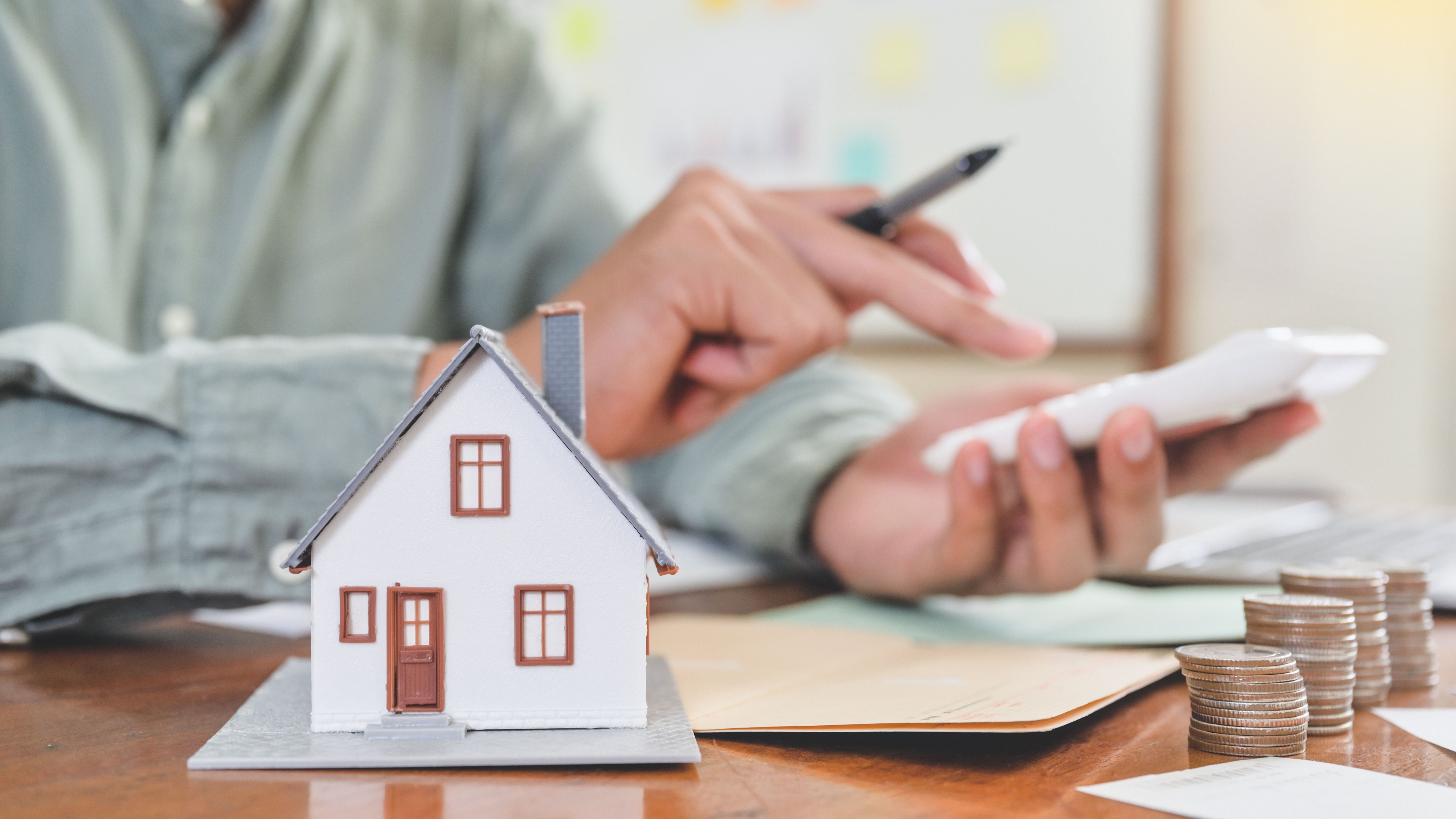 A tenant in a grey shirt holding a black pen and calculating his deposit deductions with a white calculator, next to 4 stacks of coins and a red-grey house model