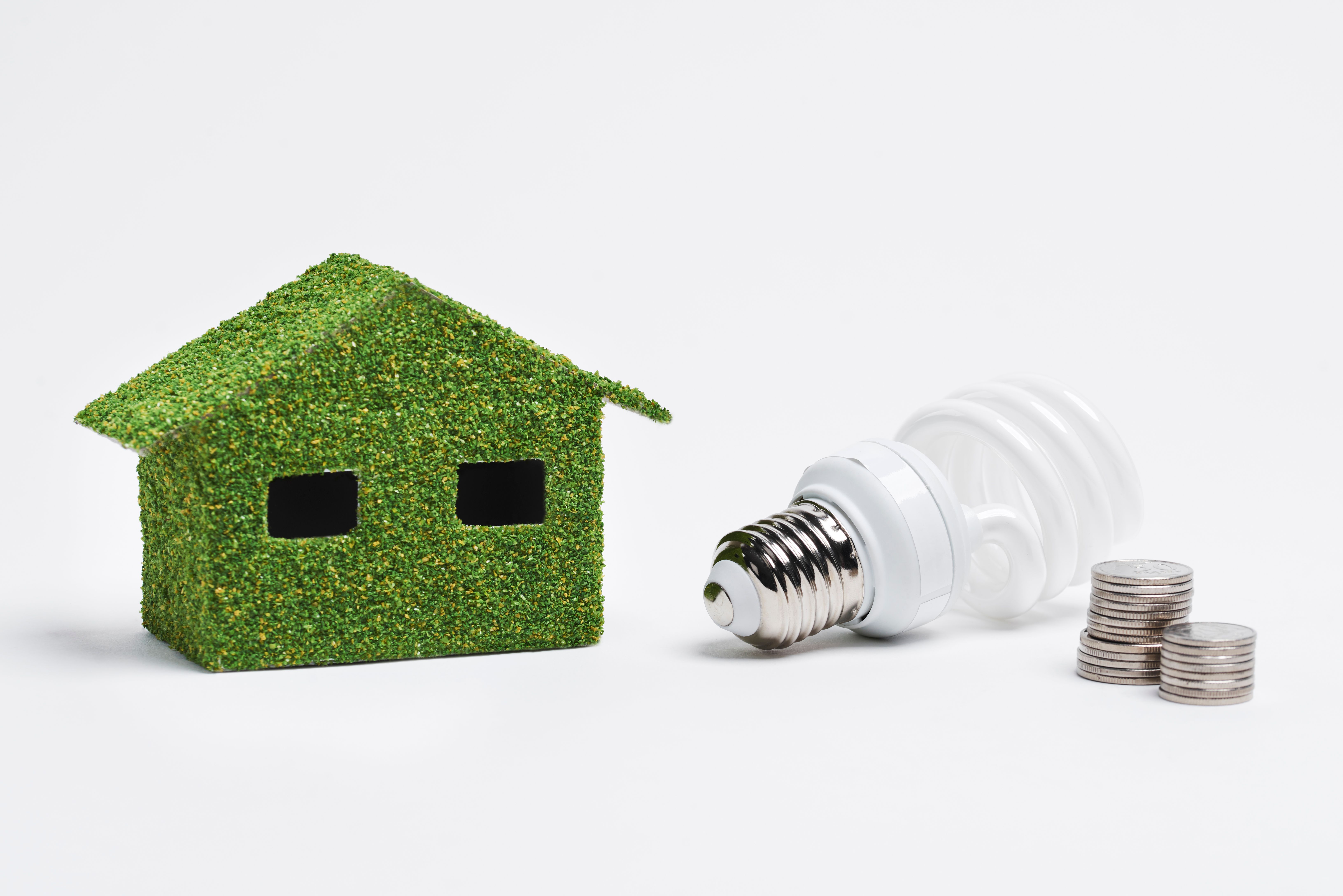 A green home model next to an energy-saving light bulb and 2 stacks of coins.