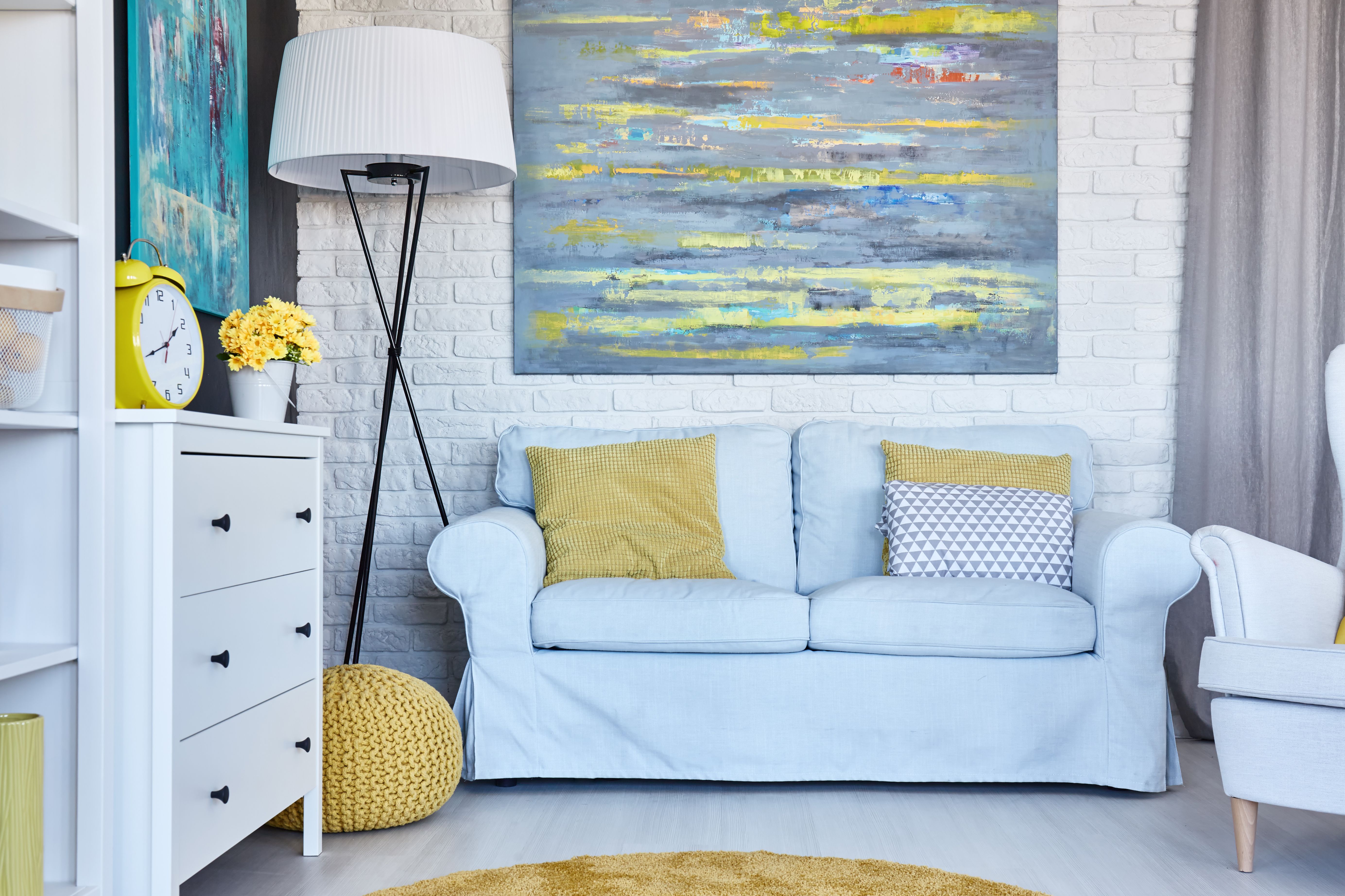  A furnished living room with a sofa, a chest of drawers, a lamp, a painting, a plant and curtains