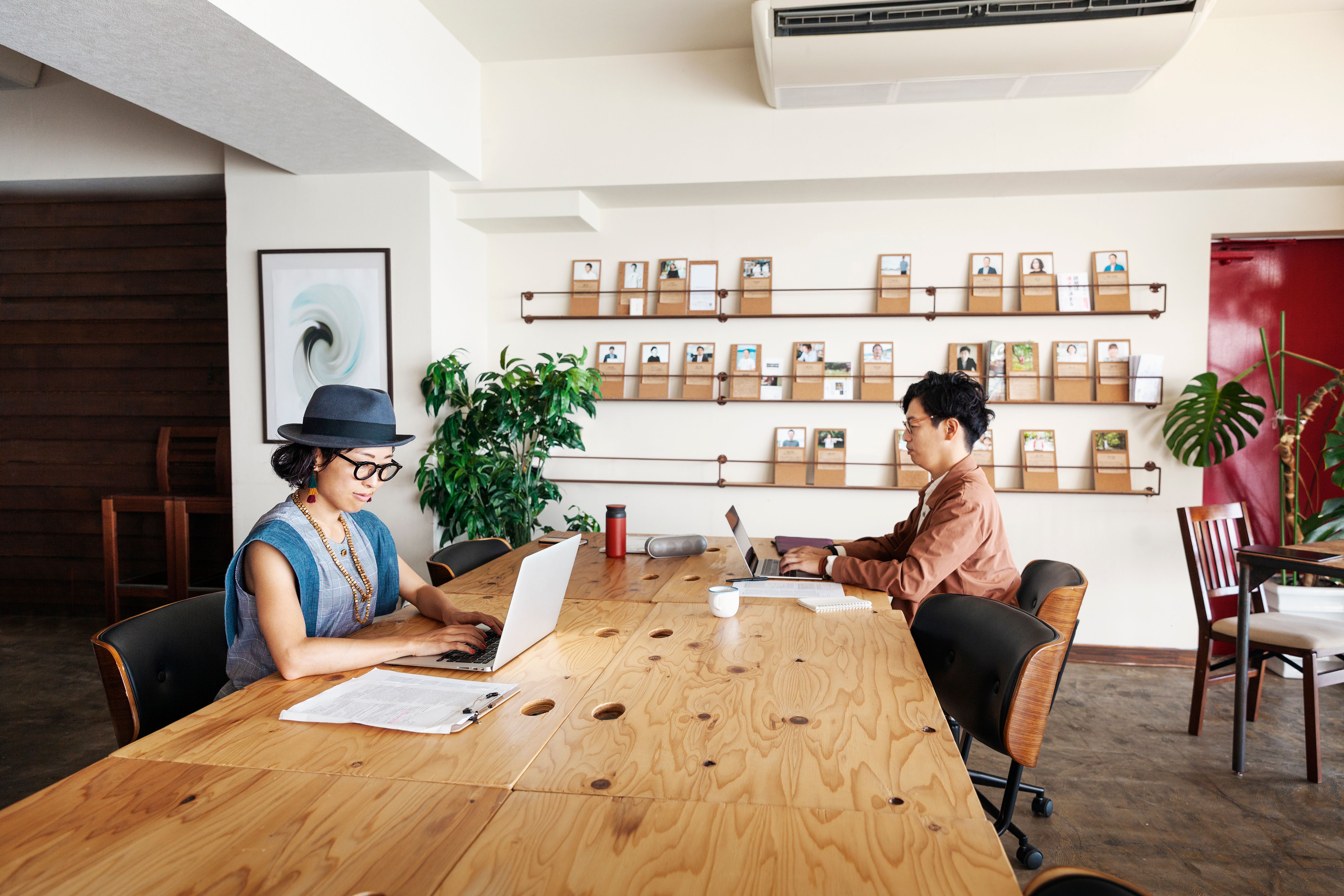 Two Japanese professionals using laptops on a desk in a co-working space with a shelf, a plant and furniture in the background