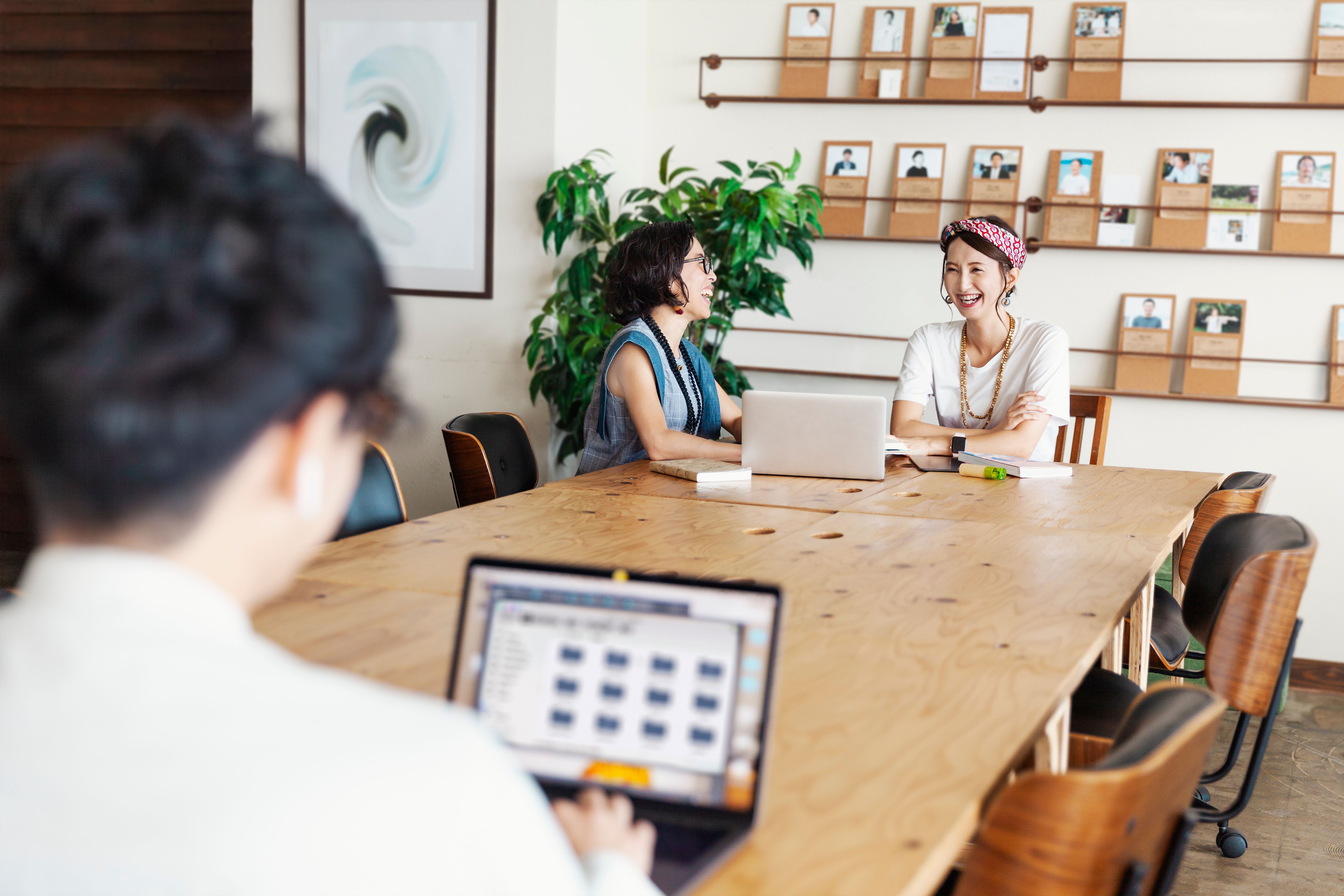 Two women having a conversation and laughing in a shared office space with a man using a laptop in the background 