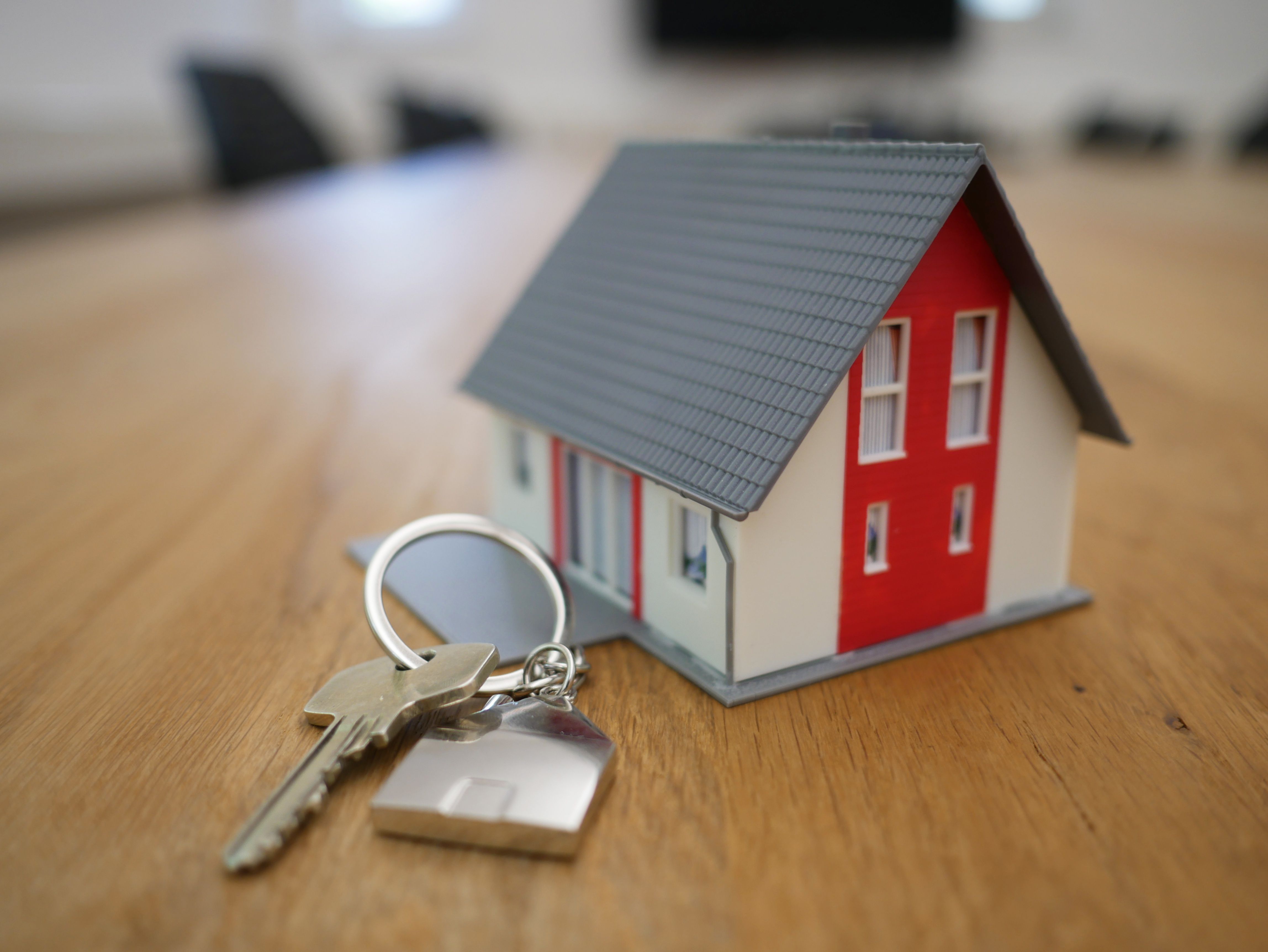 A red and grey house model next to a set of house key, both lying on a brown wooden table.