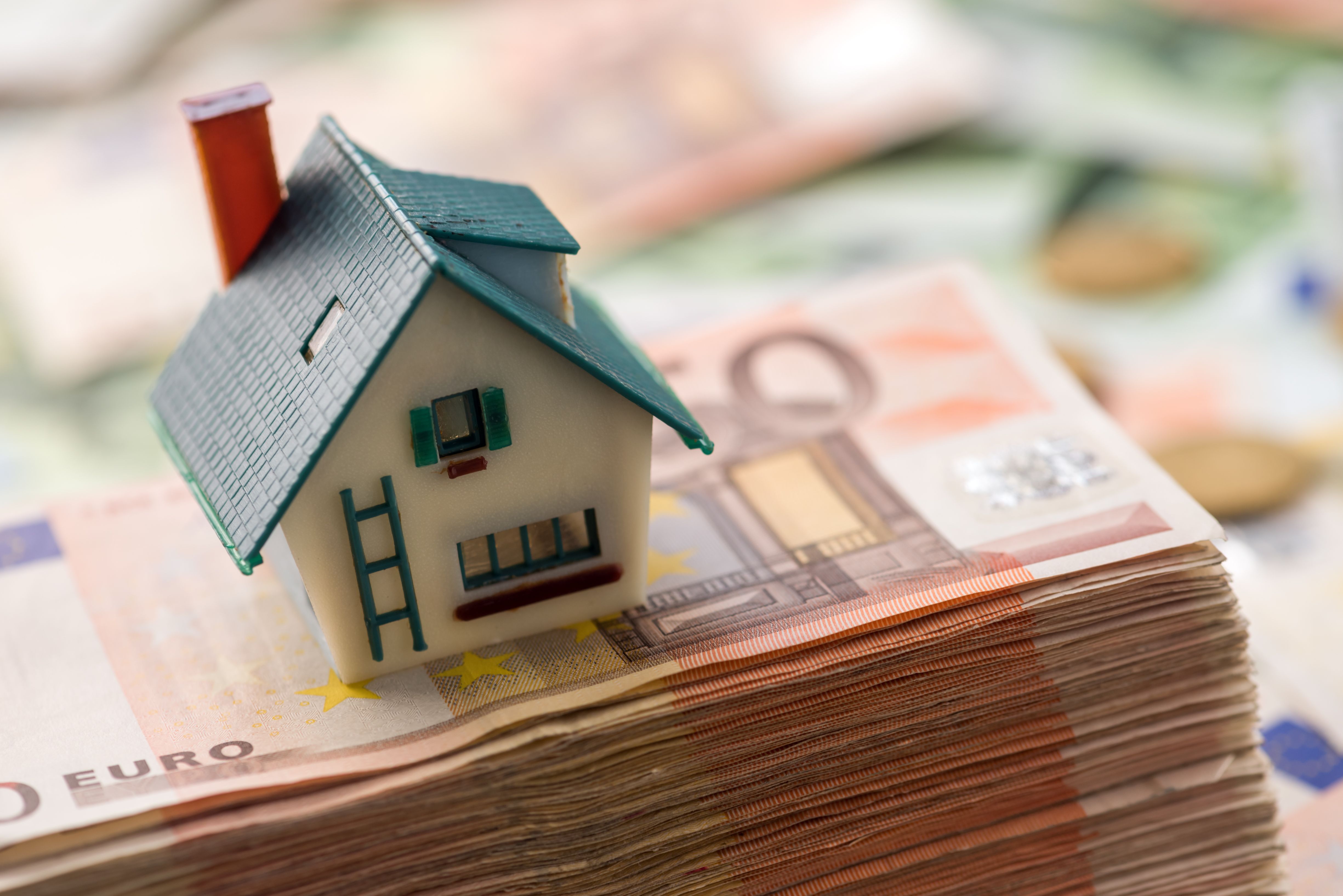 A pile of money with a model house on top against a blurred background