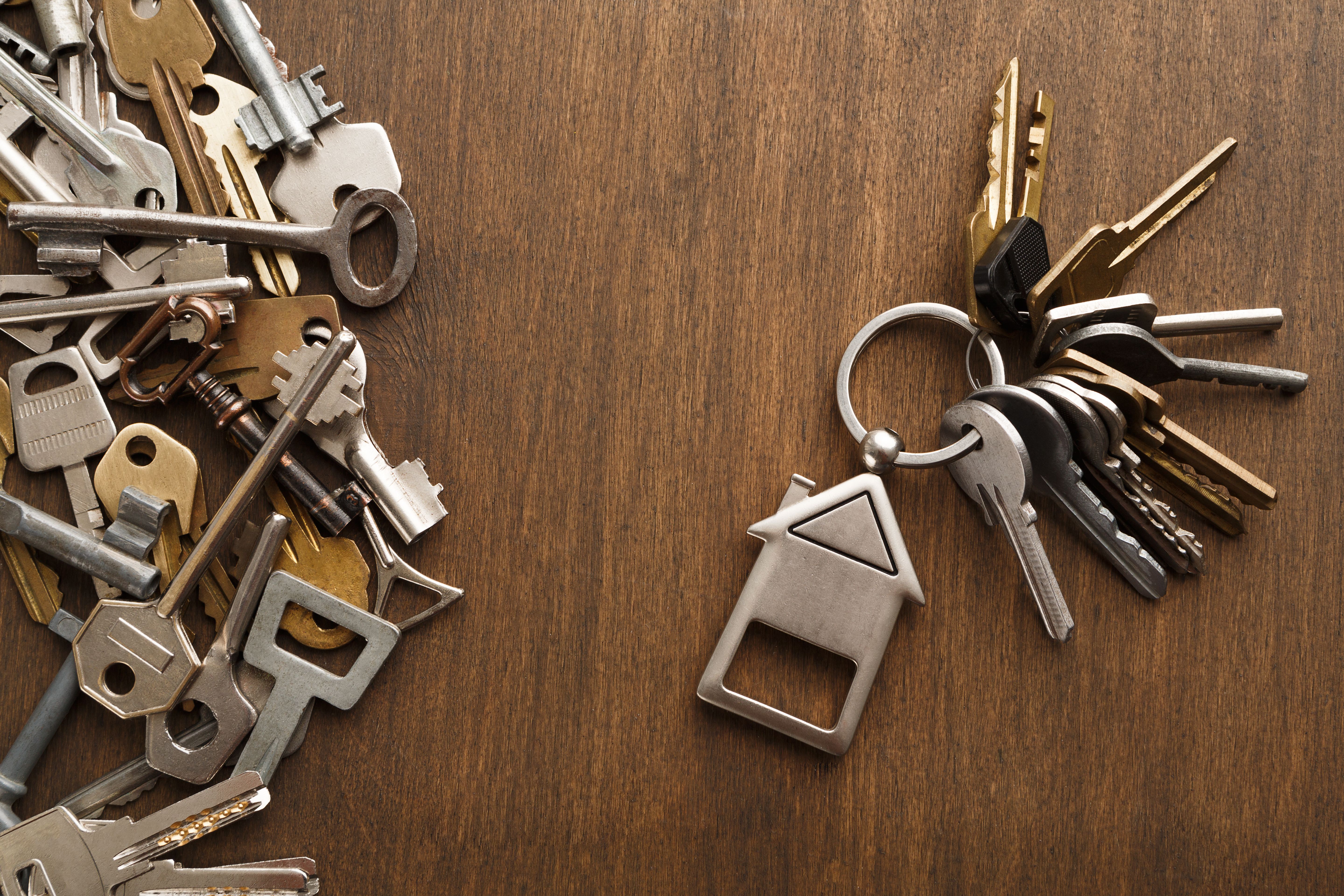 A set of 11 keys and a house model key chain next to over 20 unidentified keys on a brown wooden table.