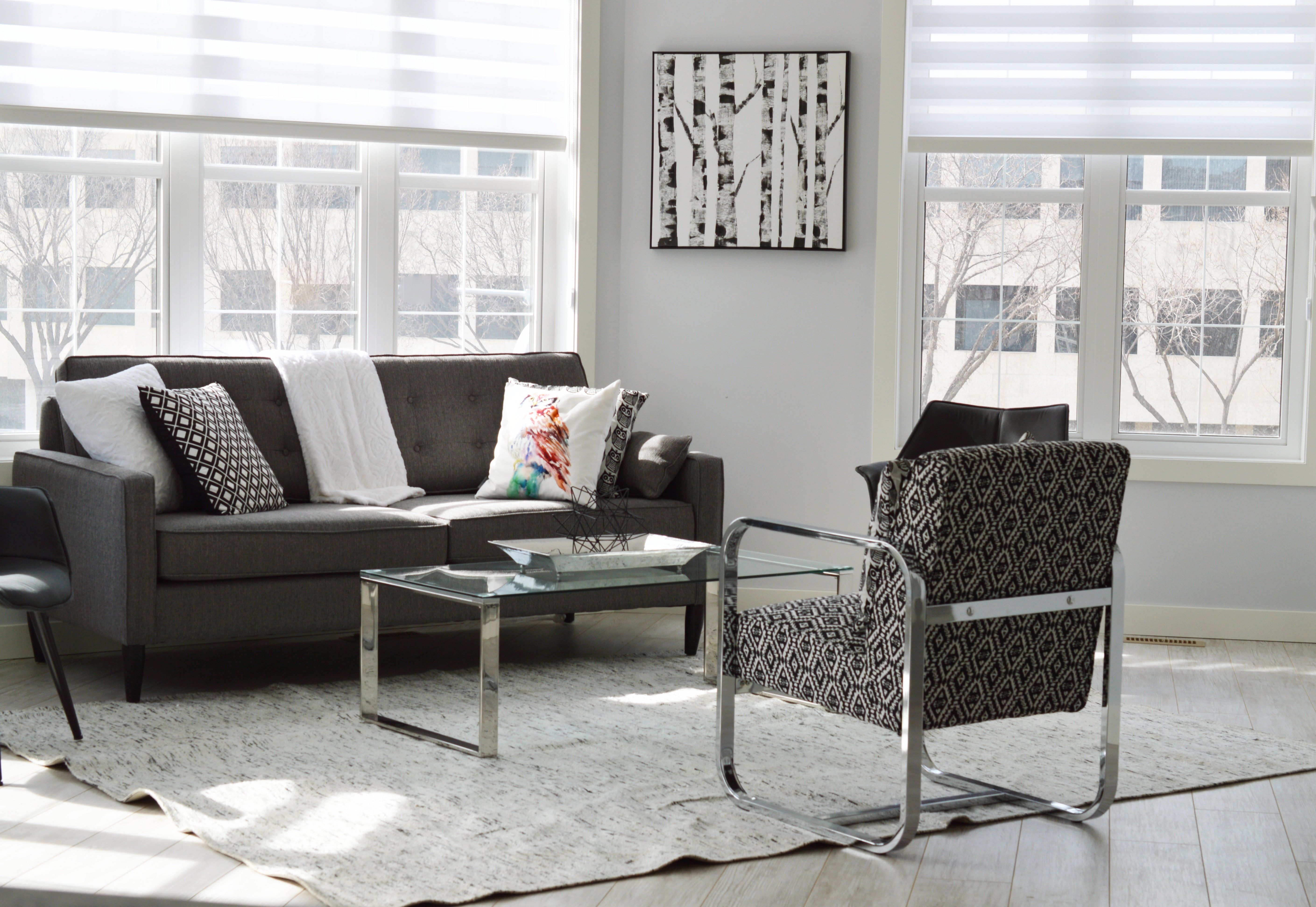 A minimalist living room with a glass coffee table, a grey sofa, an armchair, a tree painting on the wall and 2 large windows in a Build To Rent property.