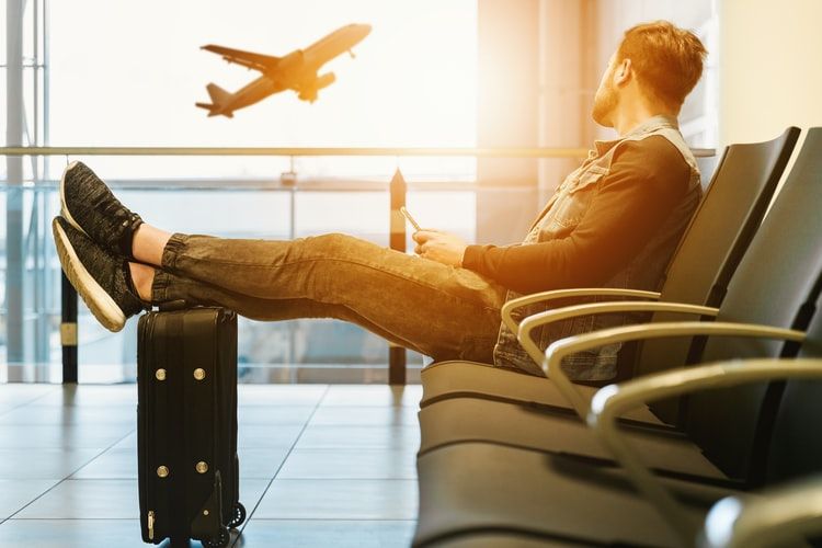 a man sitting on gang chair with feet on luggage looking on a taking off plane