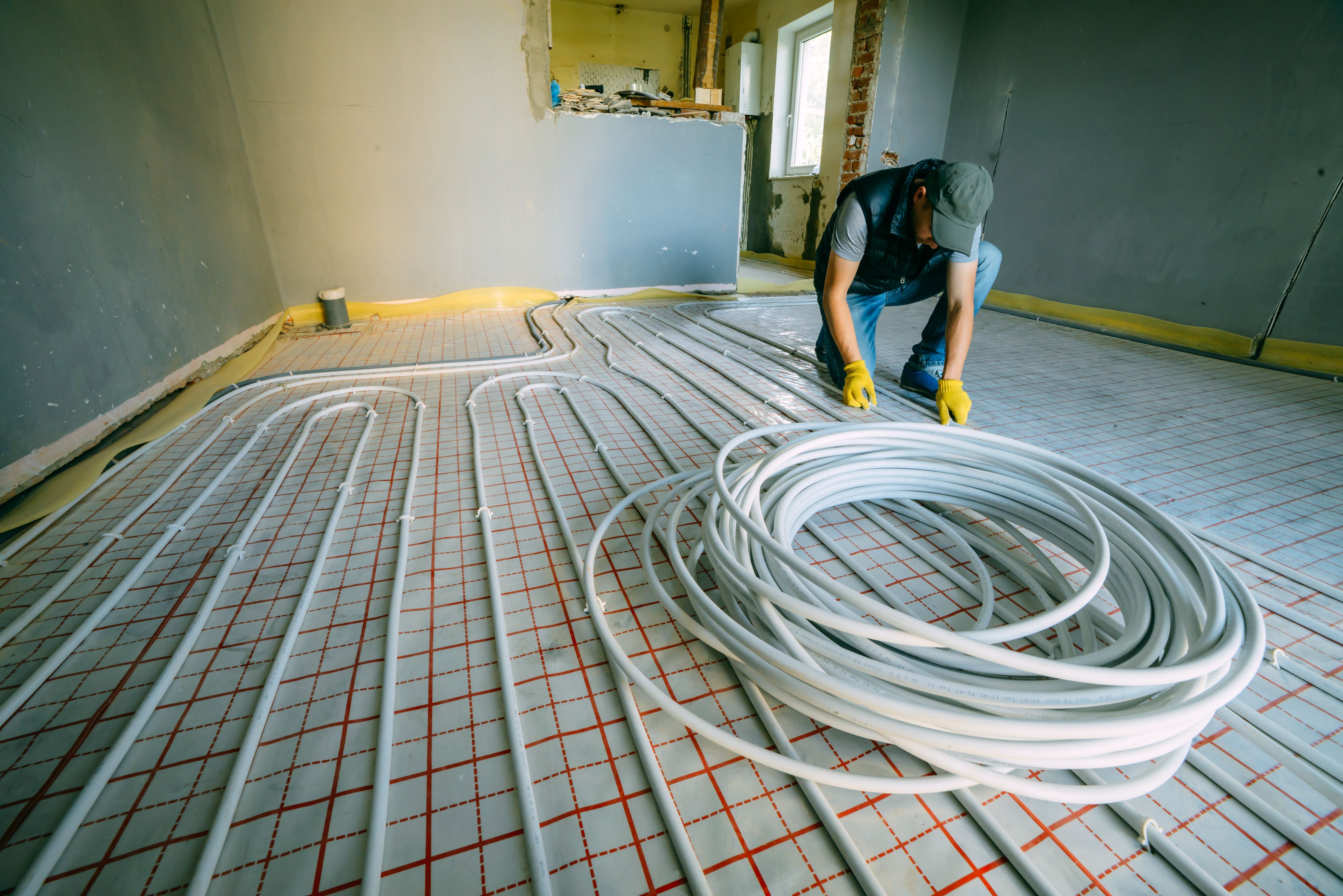 A worker installing pipes for an underfloor heating system in a room with grey walls and a doorway under construction
.jpg