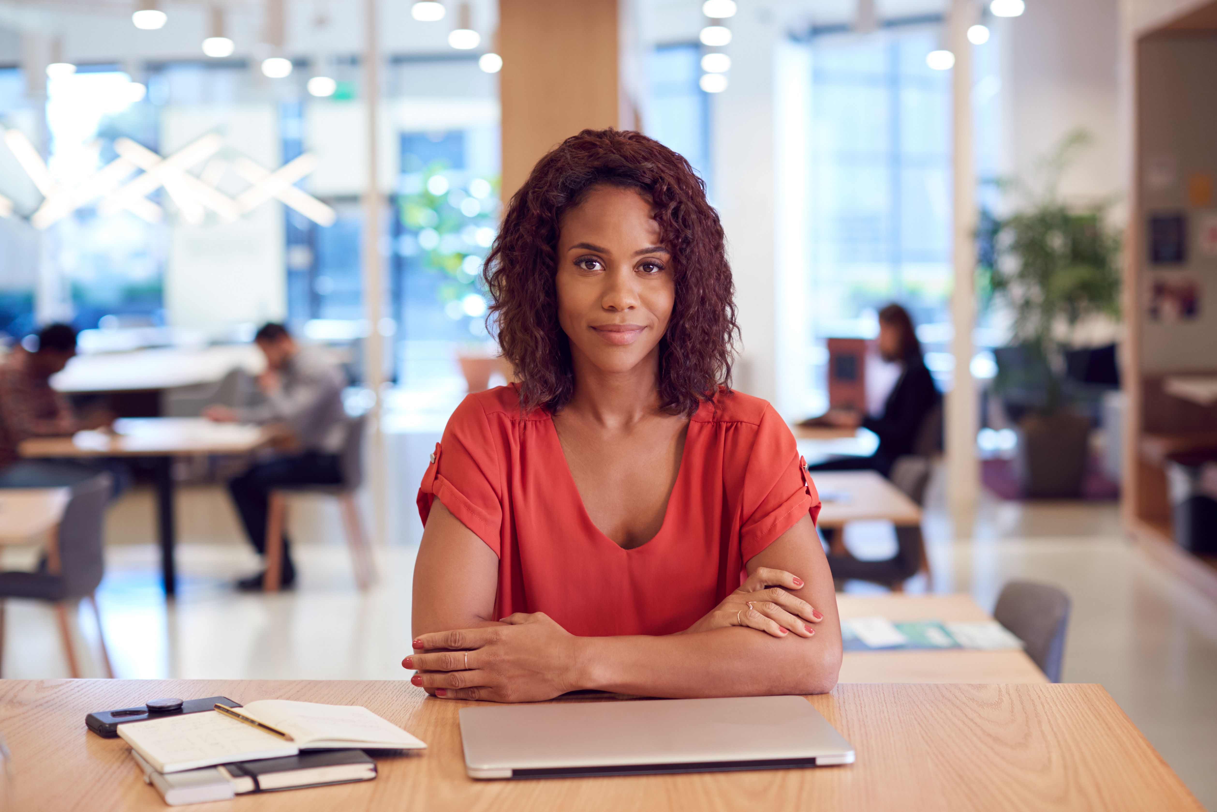 A smiling businesswoman at a work desk in a co-working space with other people and plants in the background