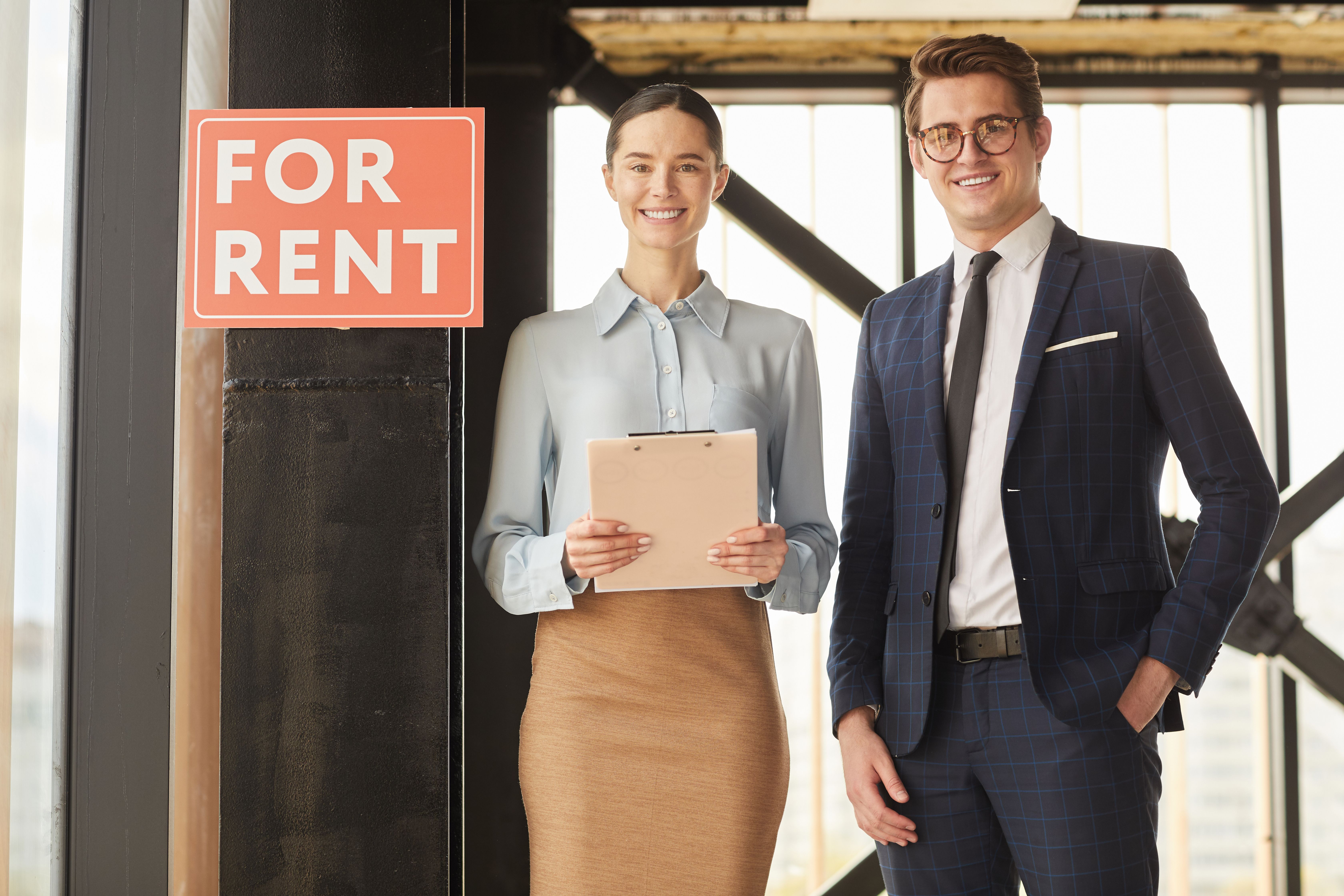 Waist up portrait of two real estate agents smiling at the camera while standing next to a red sign says For Rent
