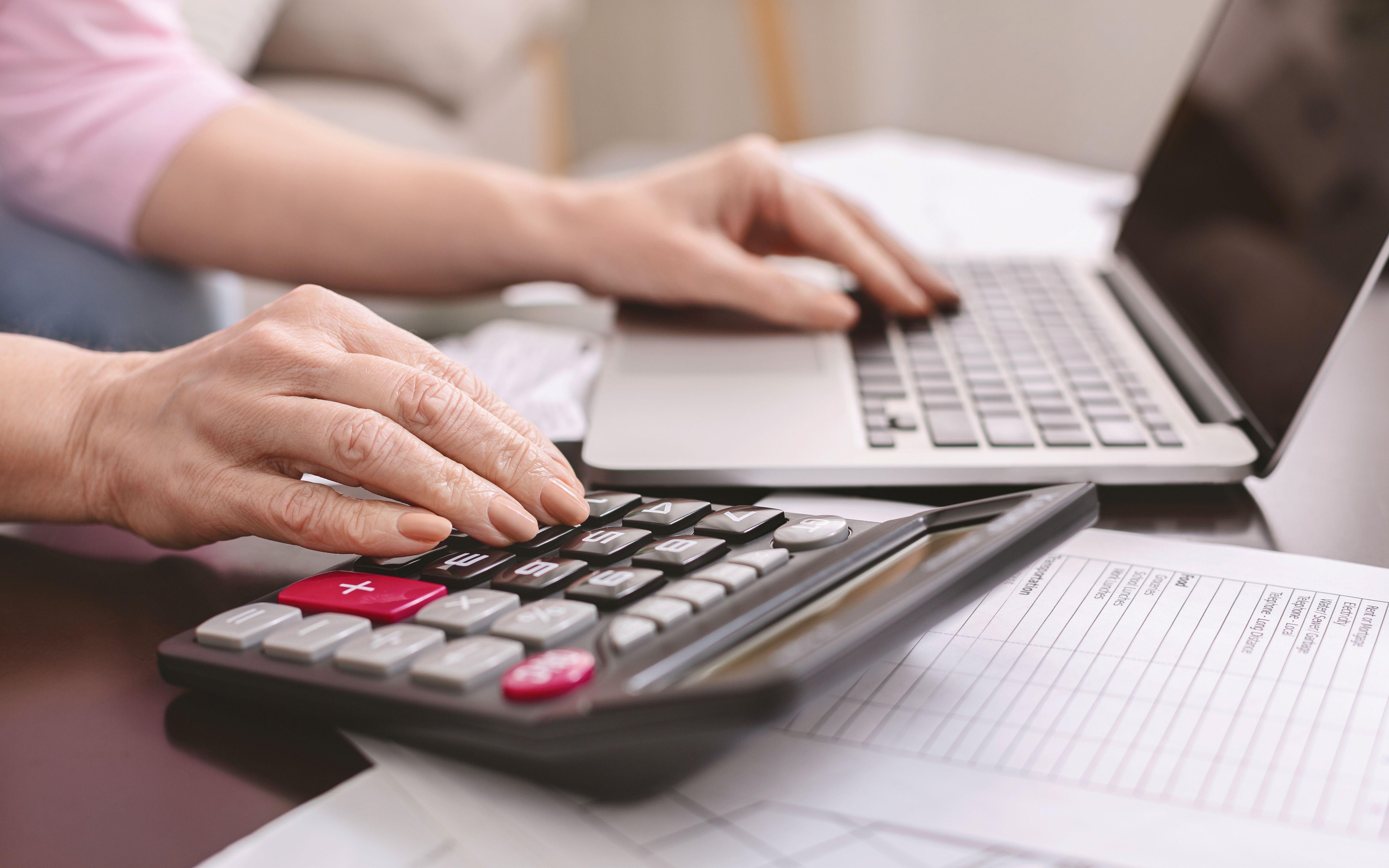  A woman reviewing documents and using a calculator and laptop on a desk against a blurred background