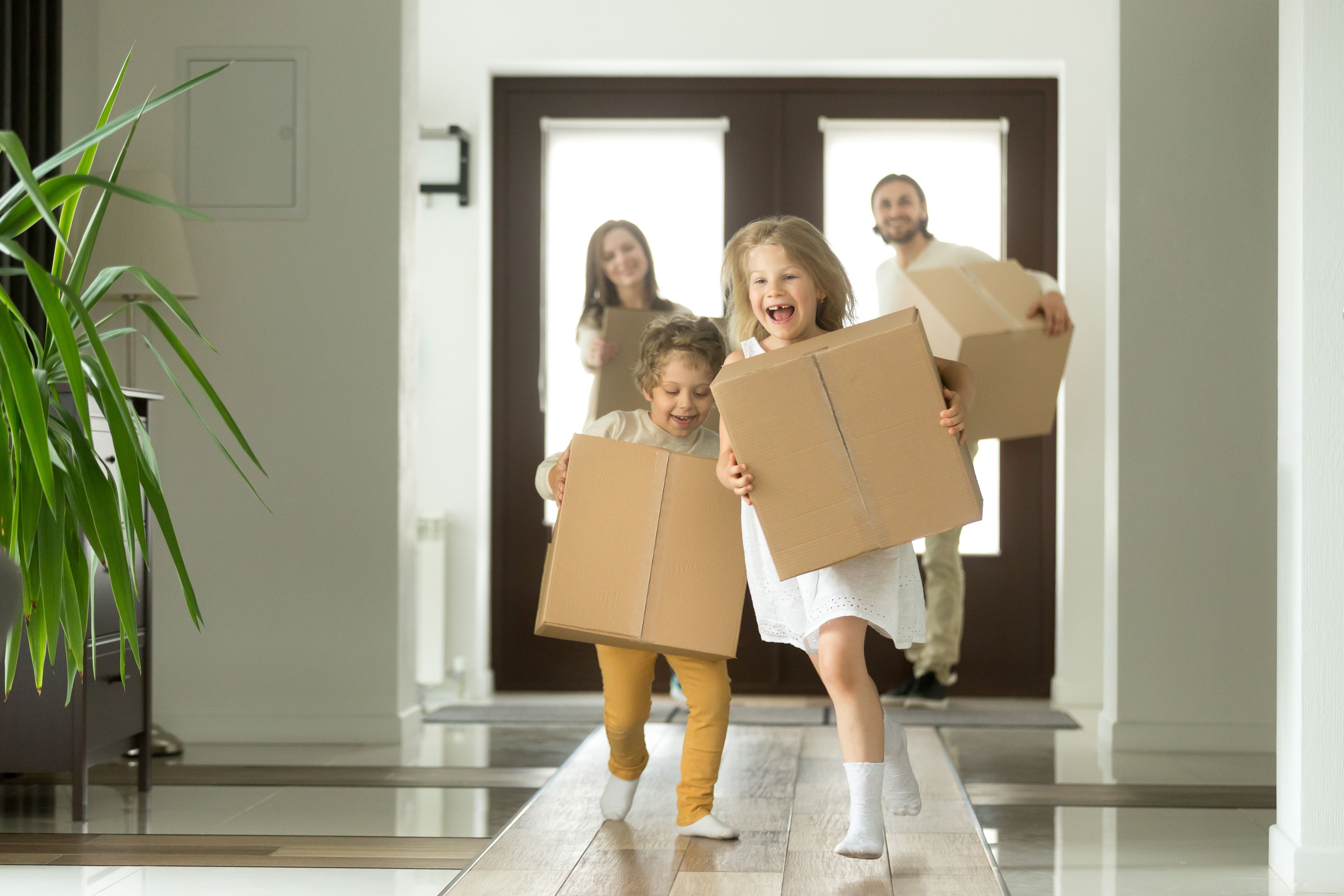 A family of 4 in white clothes holding moving boxes in their arms, running with big smiles into their new Build To Rent homes.