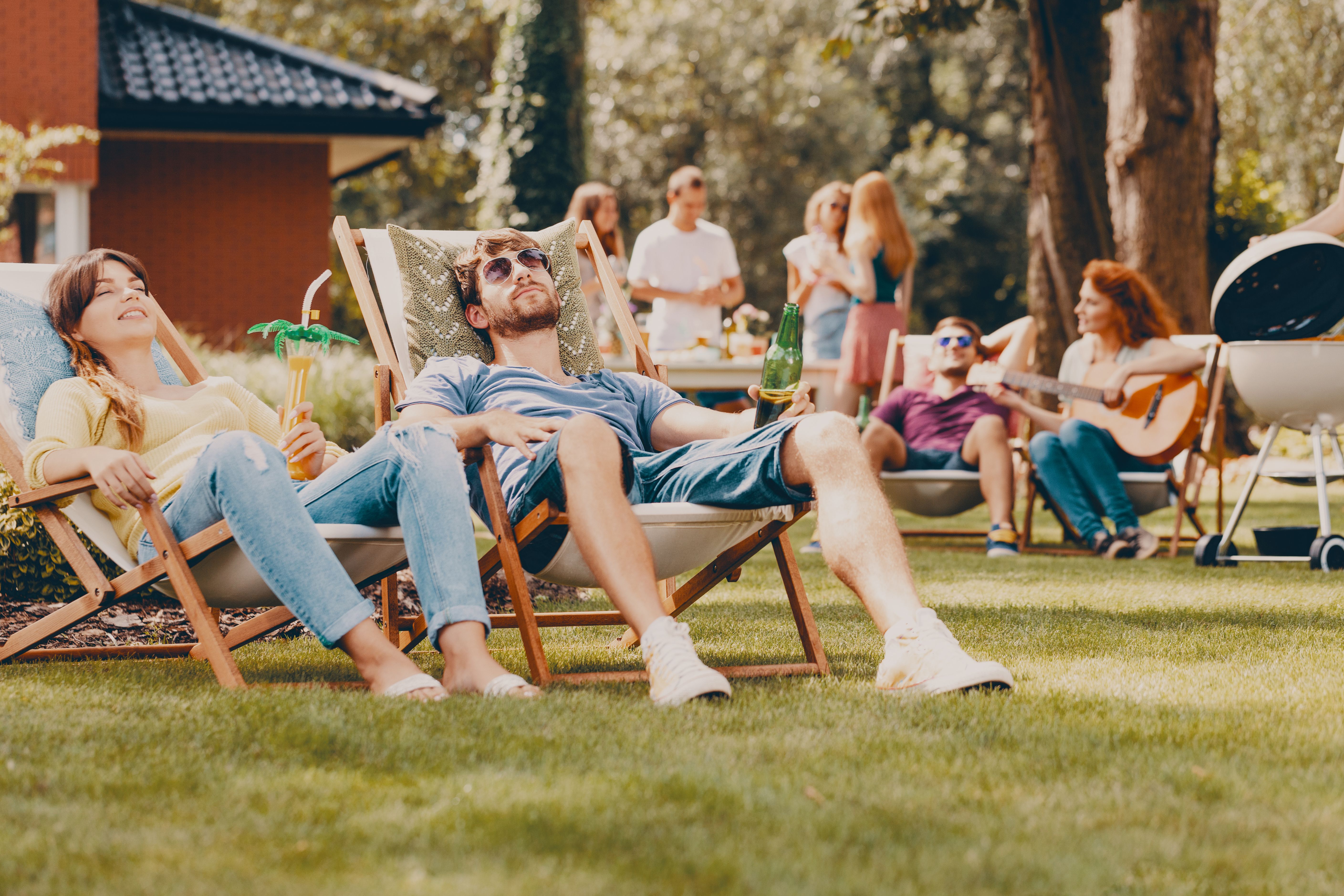 A man and a woman relaxing on sun beds during an outdoor BBQ party with other people in the background 