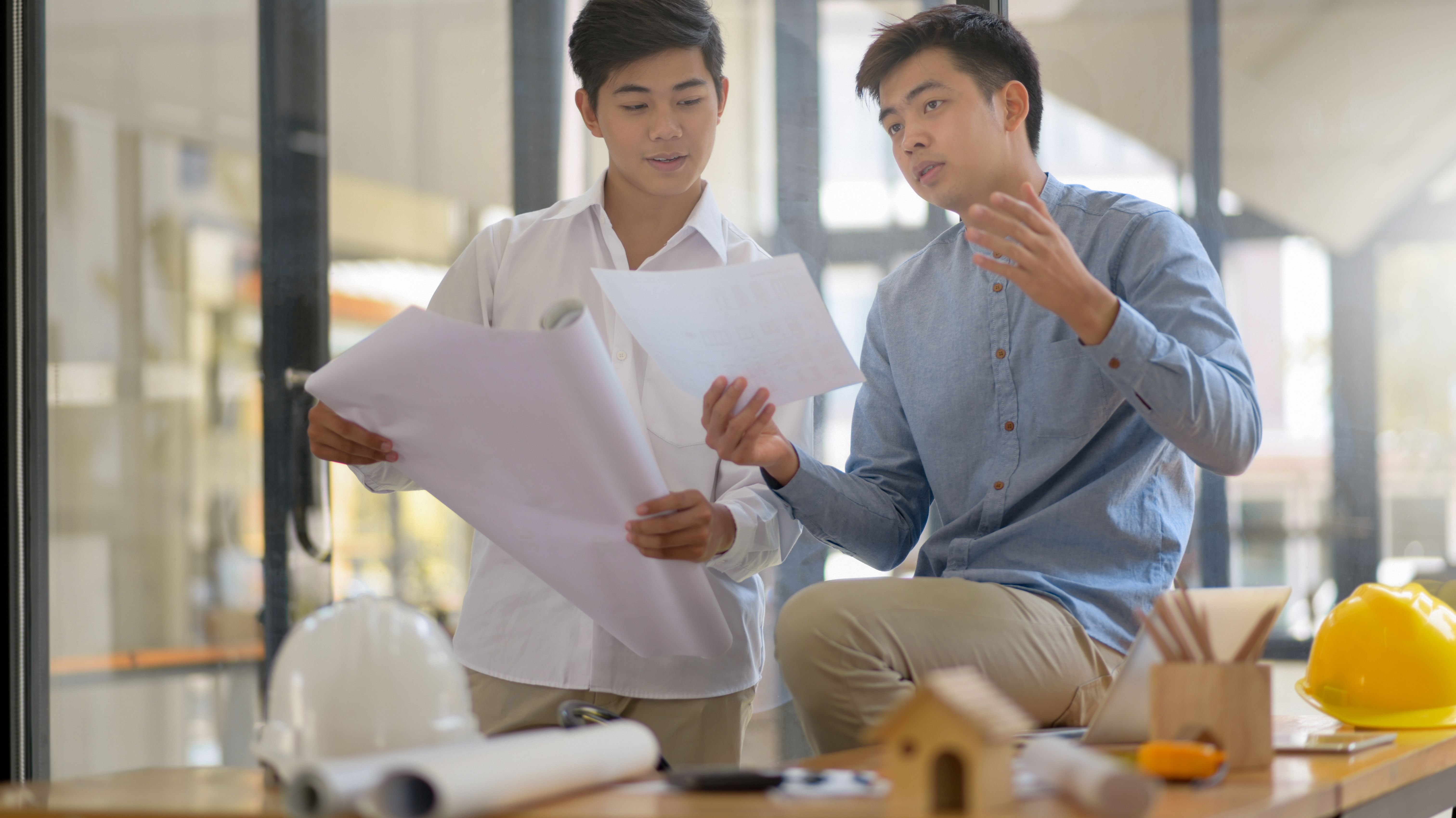 Two men holding documents and building plans having a discussion with model houses, pencils, and a construction hat on the table