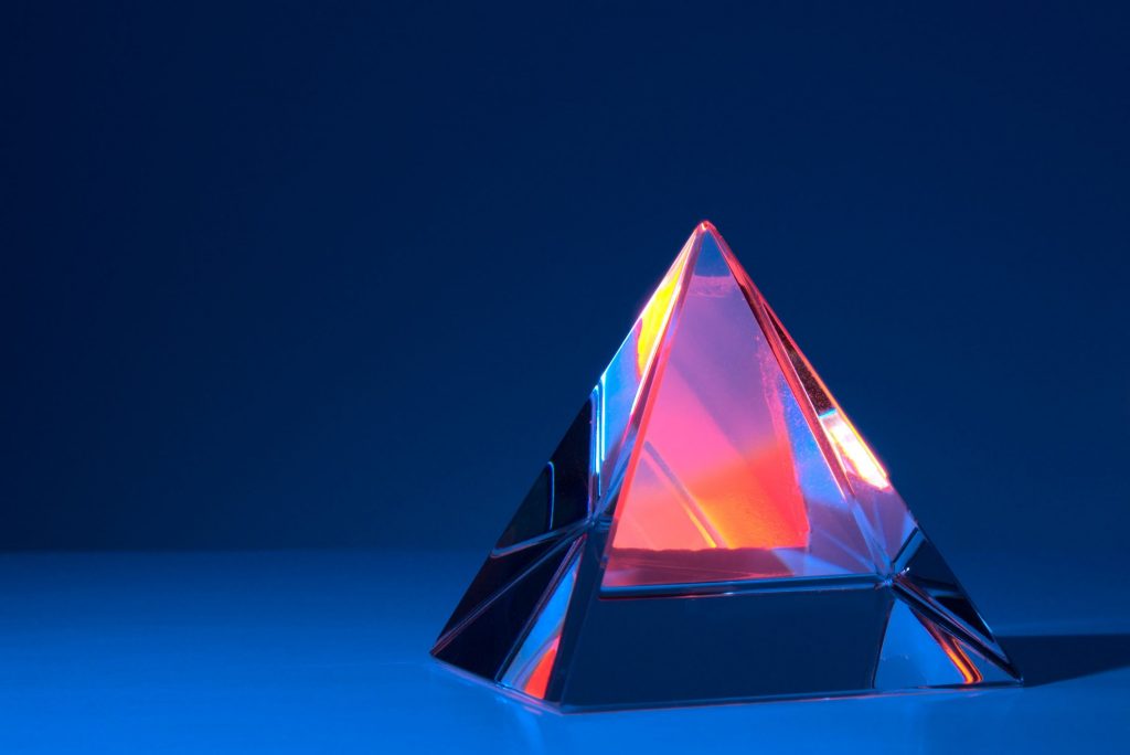  A 3D pyramid made of glass sits on a reflective surface against a dark blue background. (The image represents the search query 'Piramida skema Ponzi'.)