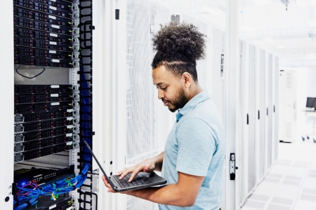 A goateed man with medium-dark skin and curly dark hair pulled into a top ponytail, wearing a short-sleeved blue shirt, stands in an IT server room looking at a laptop