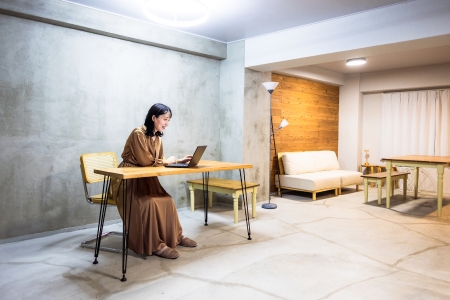 A woman with medium-length black hair sits at a bare desk in a minimalist room with a concrete wall behind her. She wears a floor-length brown dress. Beyond her is a couch with a coffee table.