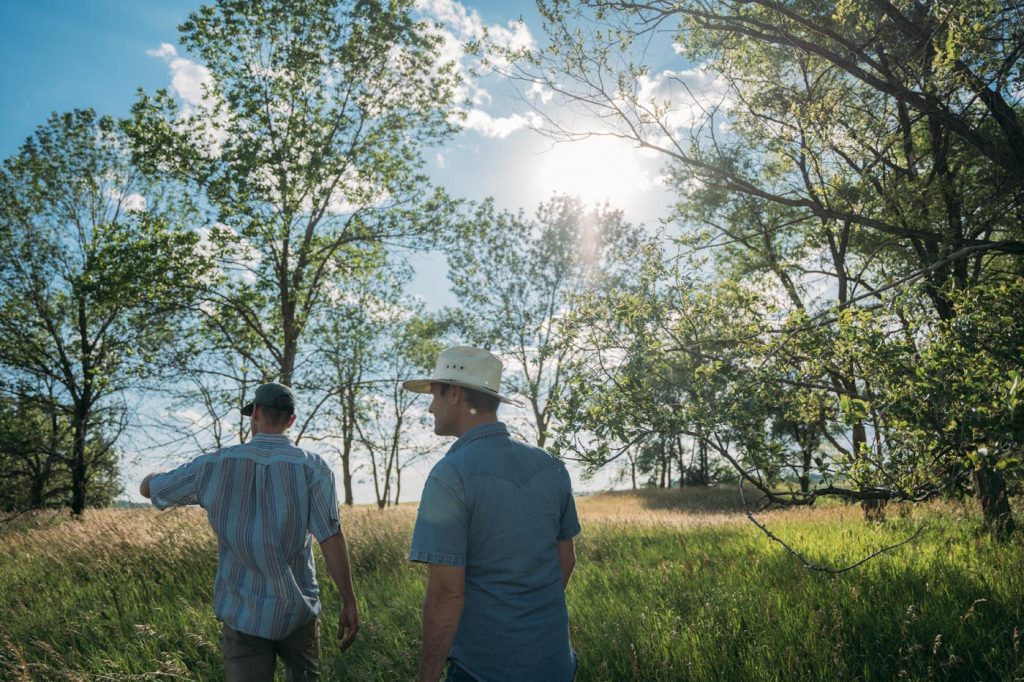 Two people wearing hats, standing in front of green grass, trees, and a blue sky with sun peaking through branches.